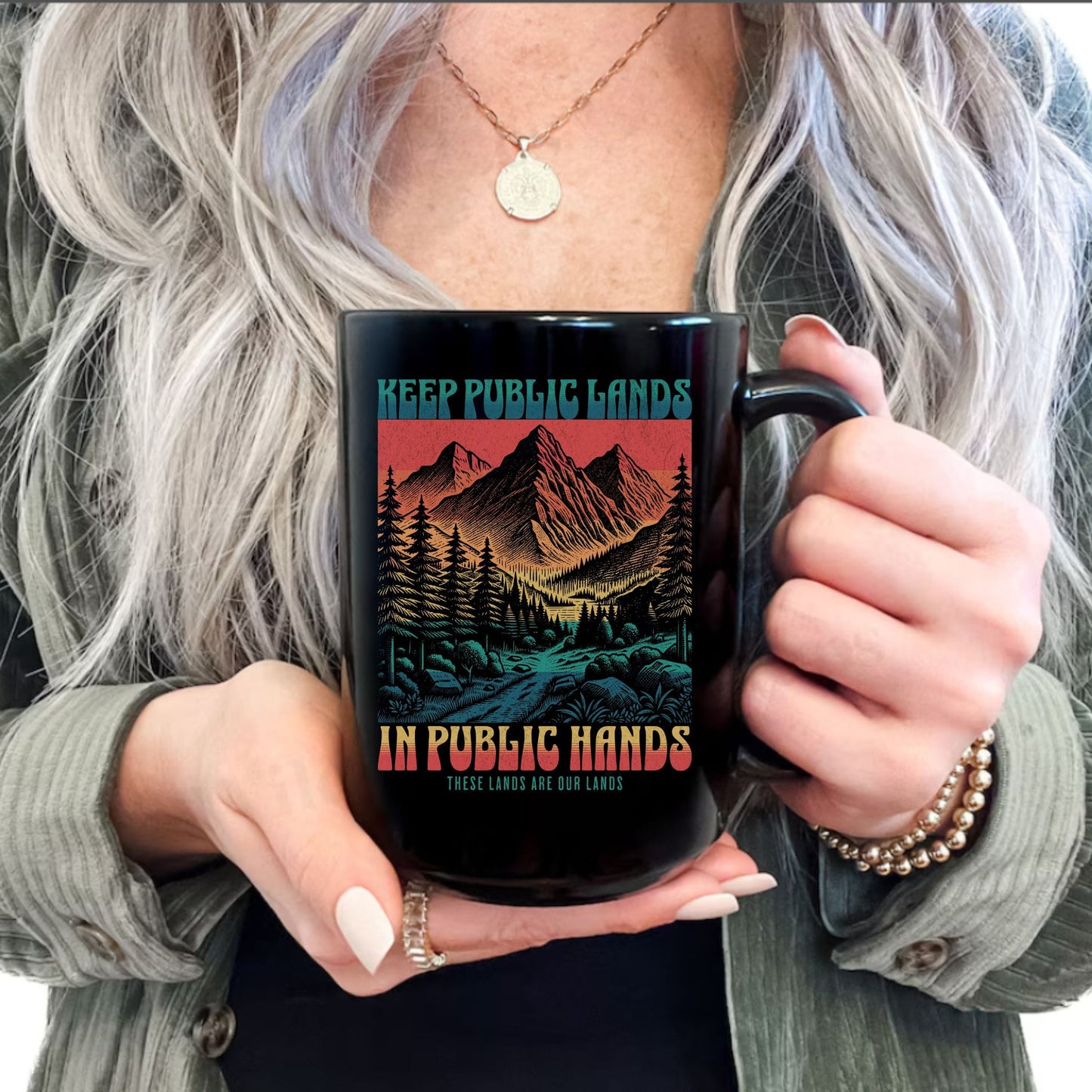 A person with long silver-gray hair holds a black ceramic mug featuring a colorful mountain landscape graphic and the text 'KEEP PUBLIC LANDS IN PUBLIC HANDS'. The person is wearing a green jacket and a gold necklace.