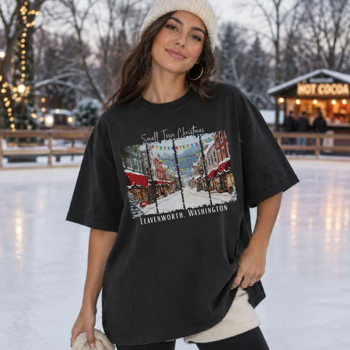 A woman wearing an oversized black graphic t-shirt with a vintage-style 'Small Town Christmas' print, posing on an outdoor ice rink with a hot cocoa stand in the background.
