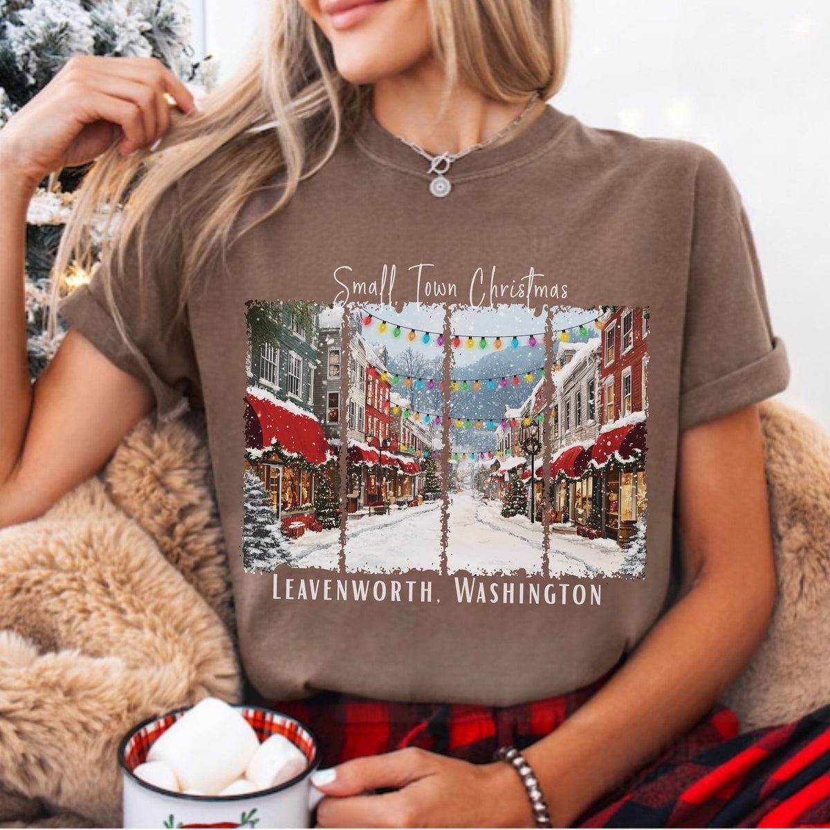 A woman wearing a brown short-sleeve graphic t-shirt with a 'Small Town Christmas' print of a snowy street in Leavenworth, Washington. She is sitting in a cozy setting with a fuzzy blanket and a mug of marshmallows.