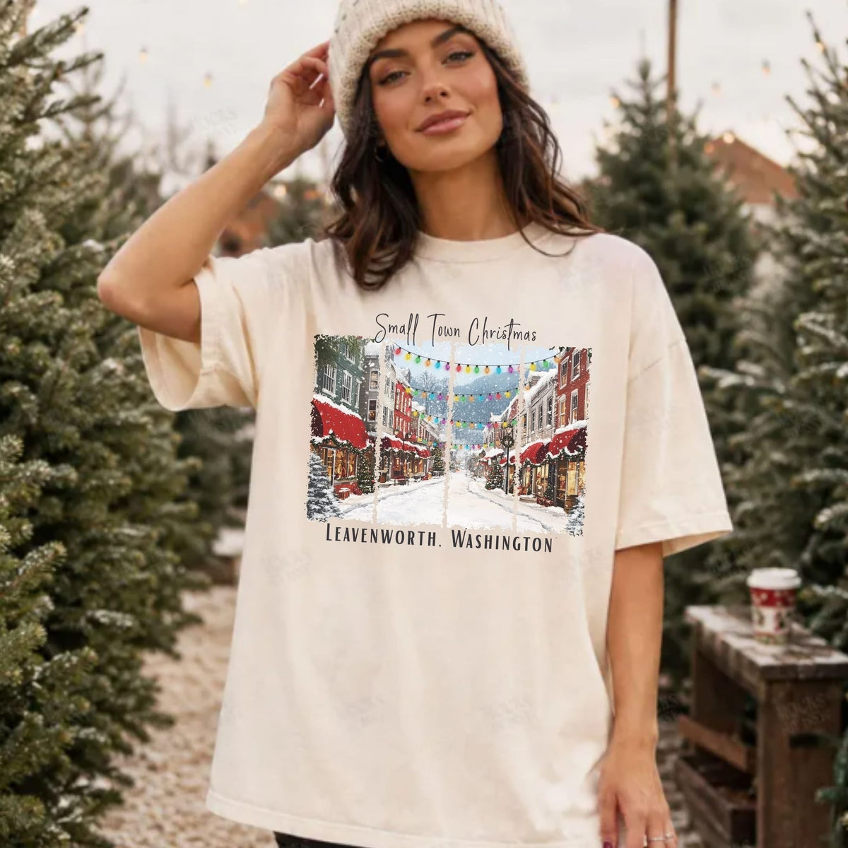 A woman wearing a cream-colored graphic t-shirt with a Christmas street scene print and a knit beanie, standing in a row of Christmas trees.