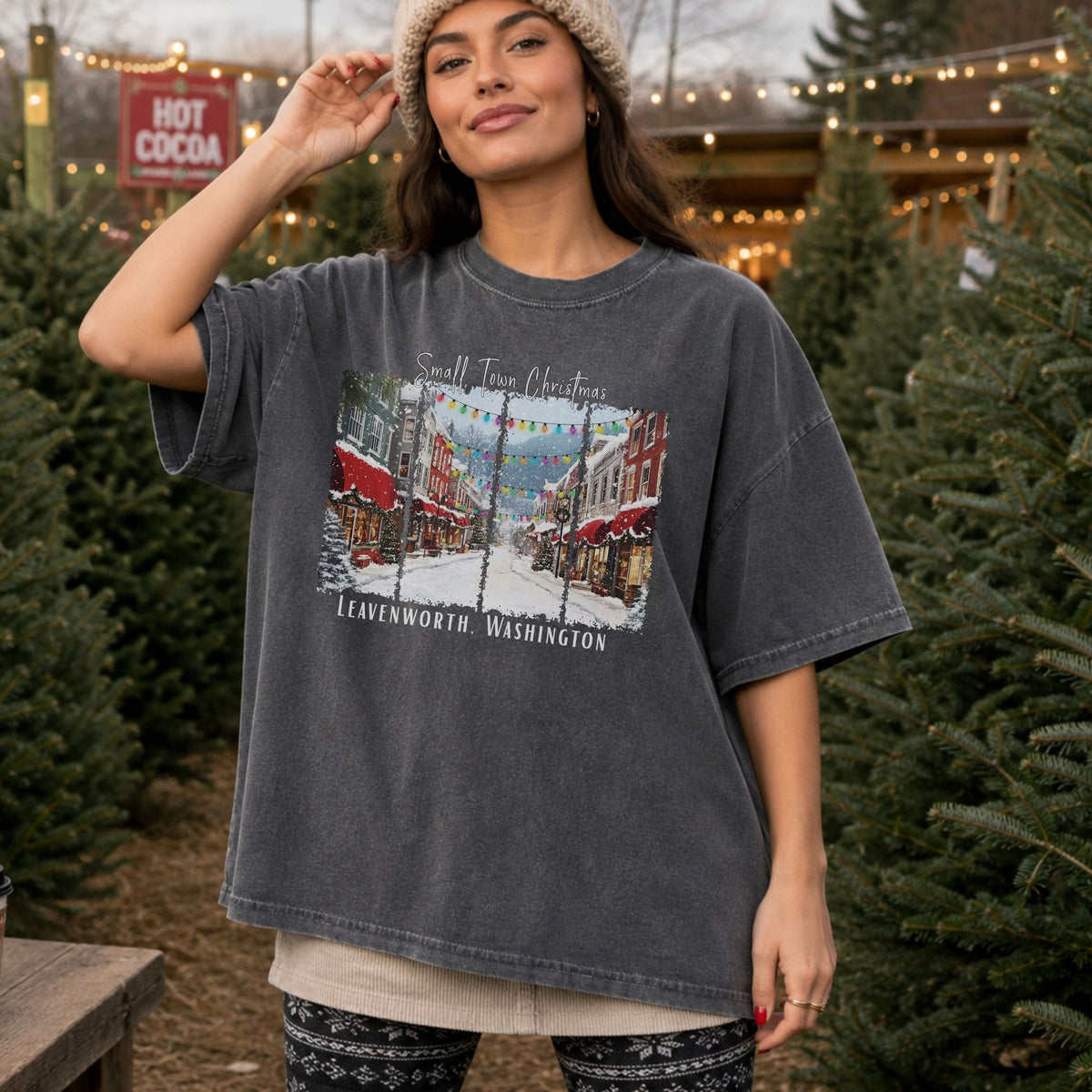 A woman wearing an oversized dark gray washed graphic t-shirt with a Christmas-themed print of 'Small Town Christmas' in Leavenworth, Washington, standing in a field of Christmas trees.