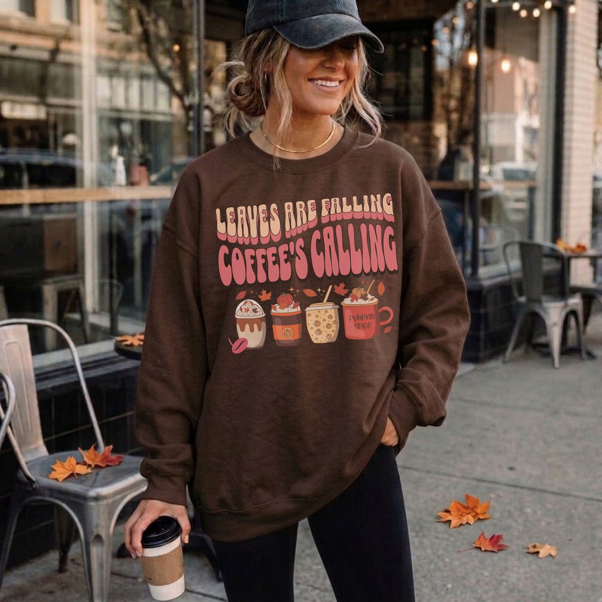 Woman wearing a dark chocolate fall coffee sweatshirt with retro autumn drinks, pumpkins, and Leaves Are Falling Coffee’s Calling design