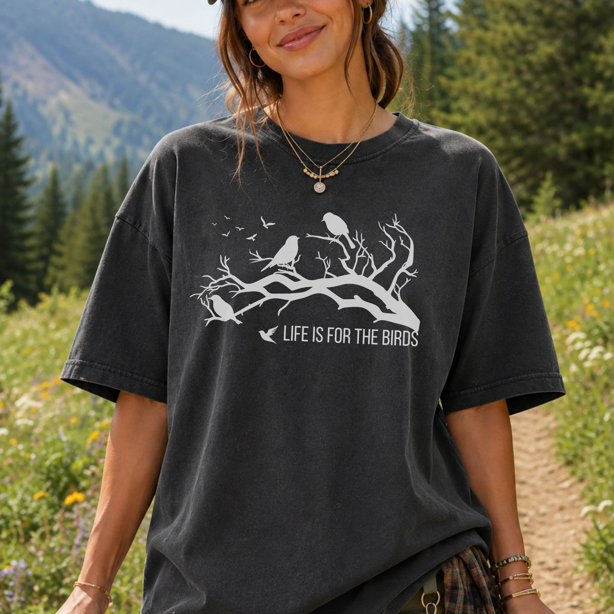 A woman wearing a faded black graphic t-shirt with a white bird design and the text 'LIFE IS FOR THE BIRDS', set against a blurred outdoor mountain and forest background.