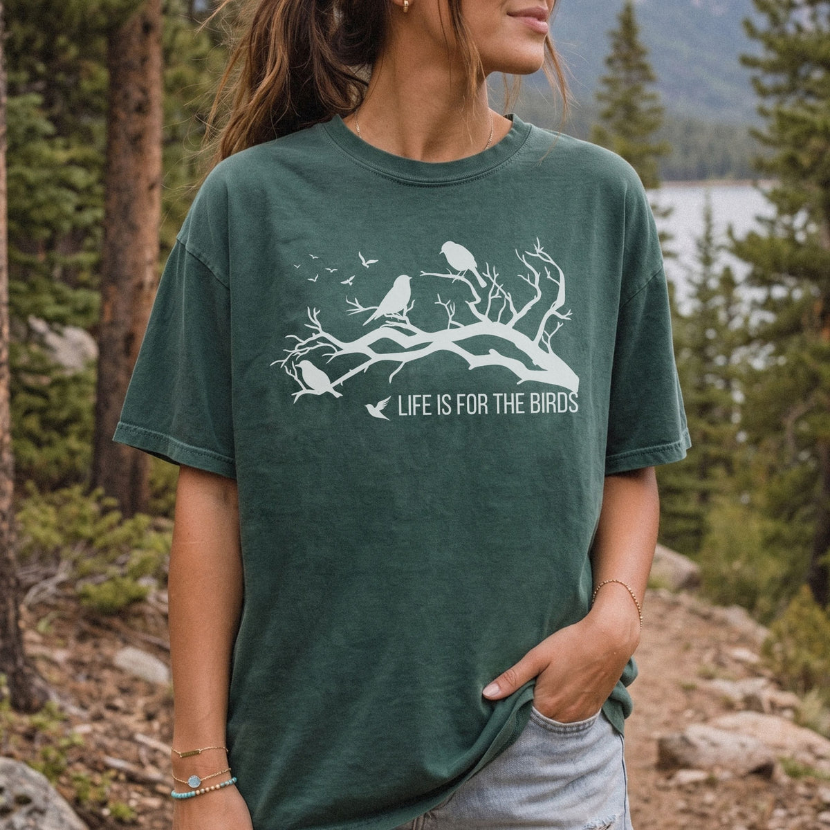 A woman wearing a dark green graphic t-shirt with a white bird design and the text 'LIFE IS FOR THE BIRDS', photographed outdoors in a forest setting.