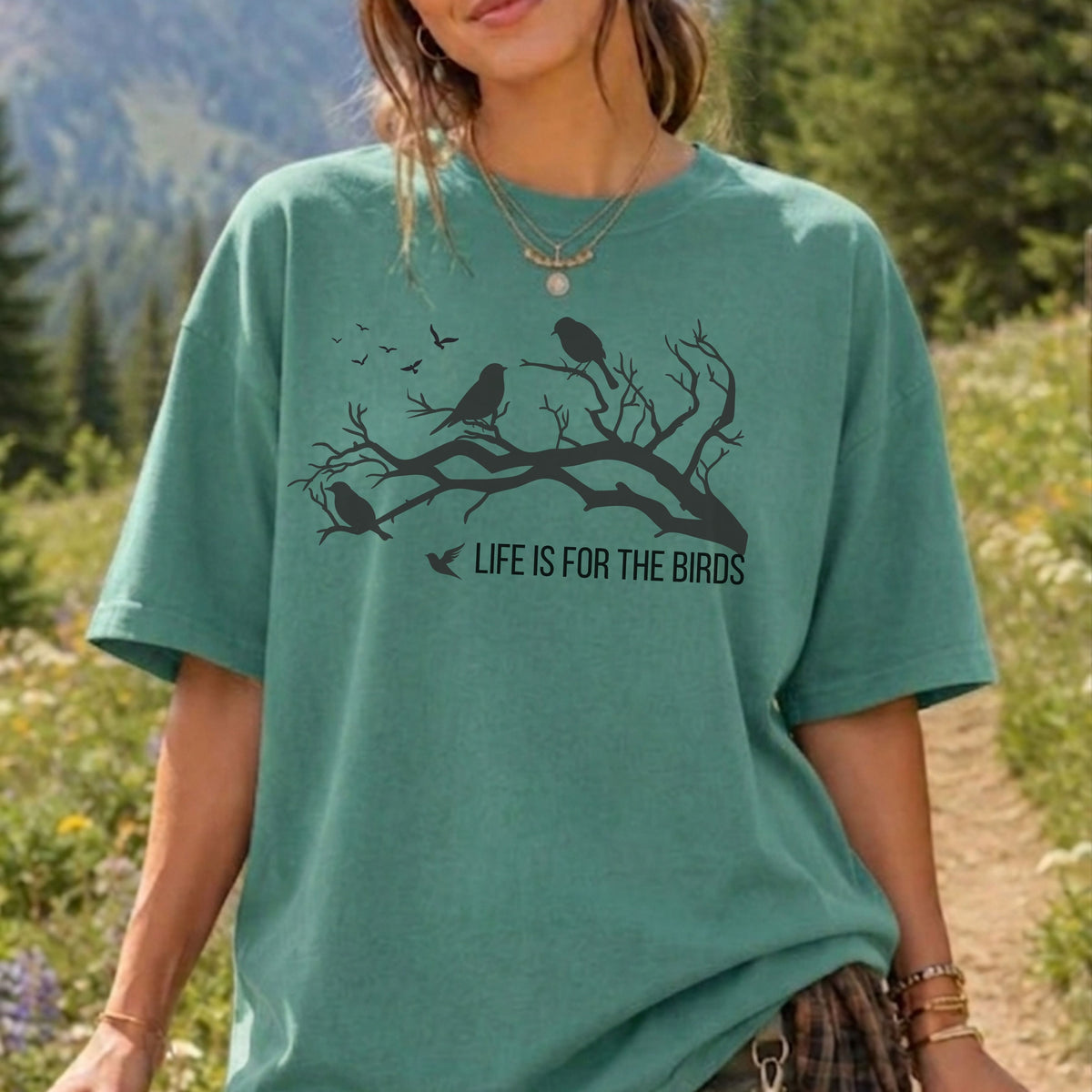 A woman wearing an oversized sage green graphic t-shirt with a black bird and branch print and the text 'LIFE IS FOR THE BIRDS'. The background is a blurred outdoor nature scene with trees and a path.