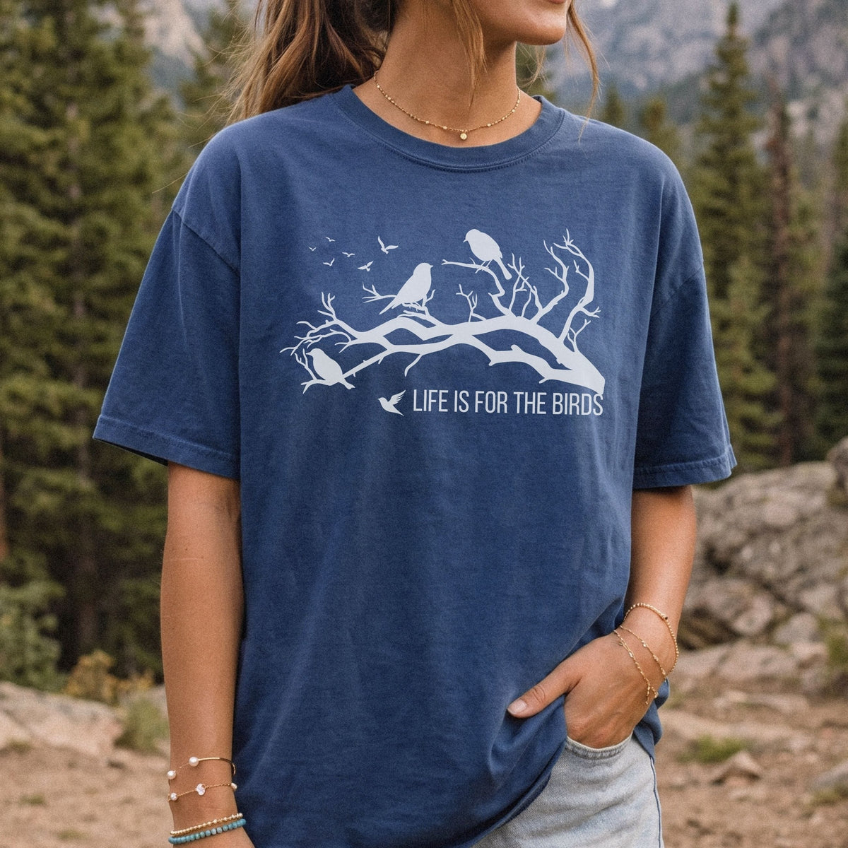 A woman wearing a navy blue graphic t-shirt with a white bird design and the text 'LIFE IS FOR THE BIRDS', set against a natural forest background.
