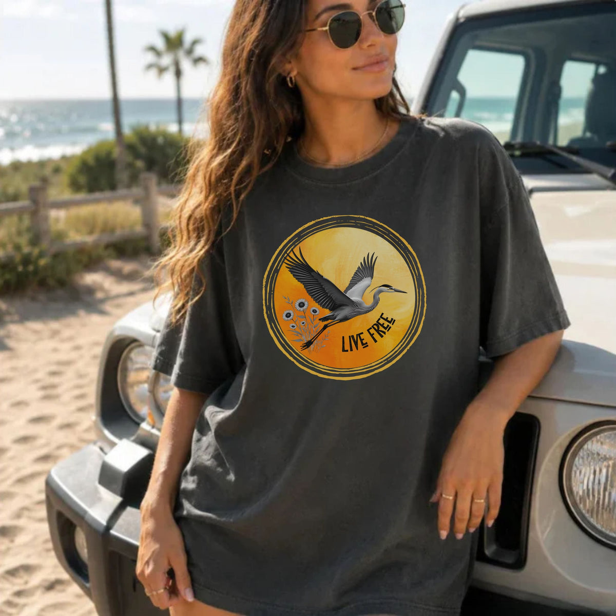 A woman wearing a dark charcoal gray graphic t-shirt with a heron and 'LIVE FREE' print, posing outdoors next to a white vehicle on a sandy beach with the ocean in the background.