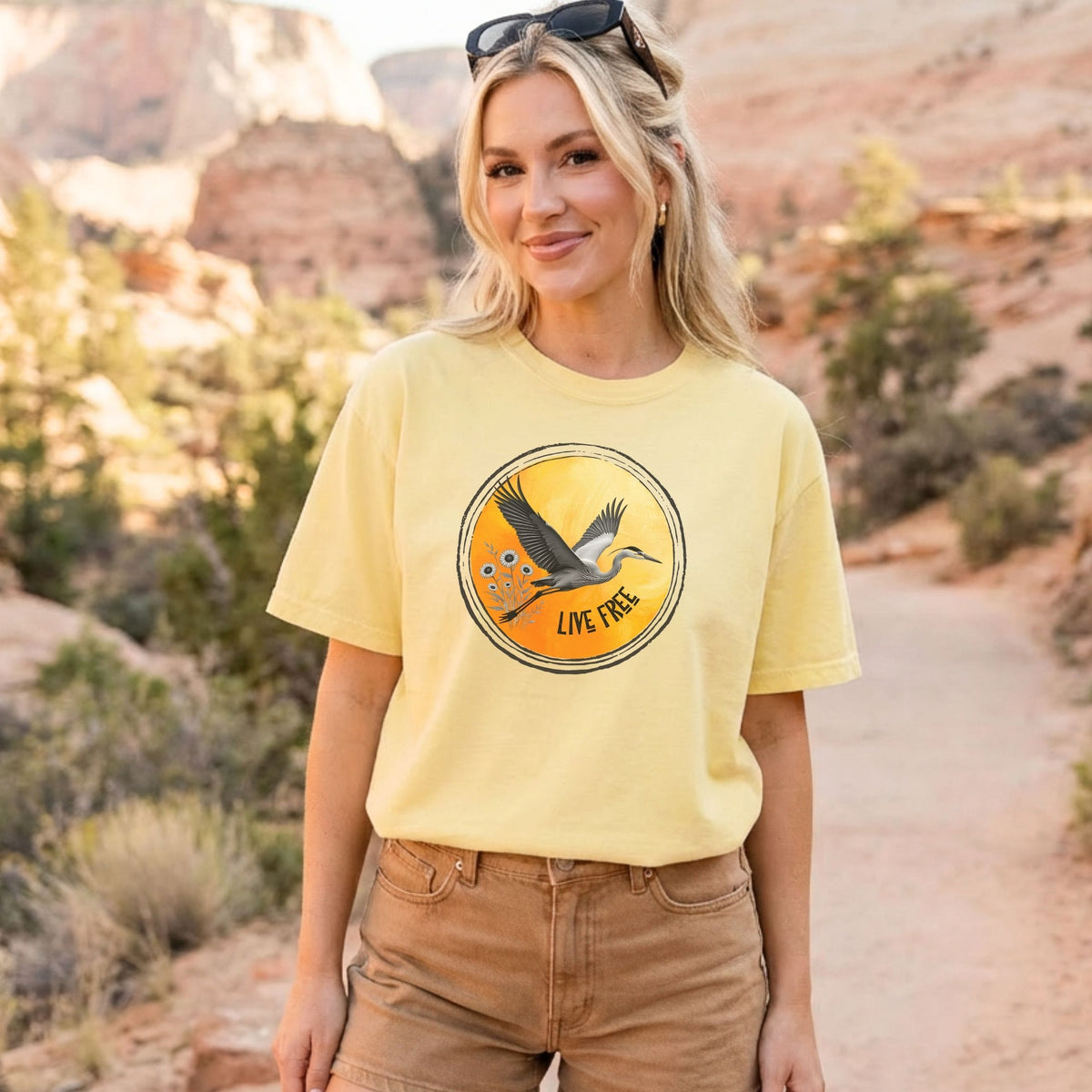 A woman wearing a pale yellow graphic t-shirt with a circular 'Live Fish' design, standing outdoors in a desert canyon setting.