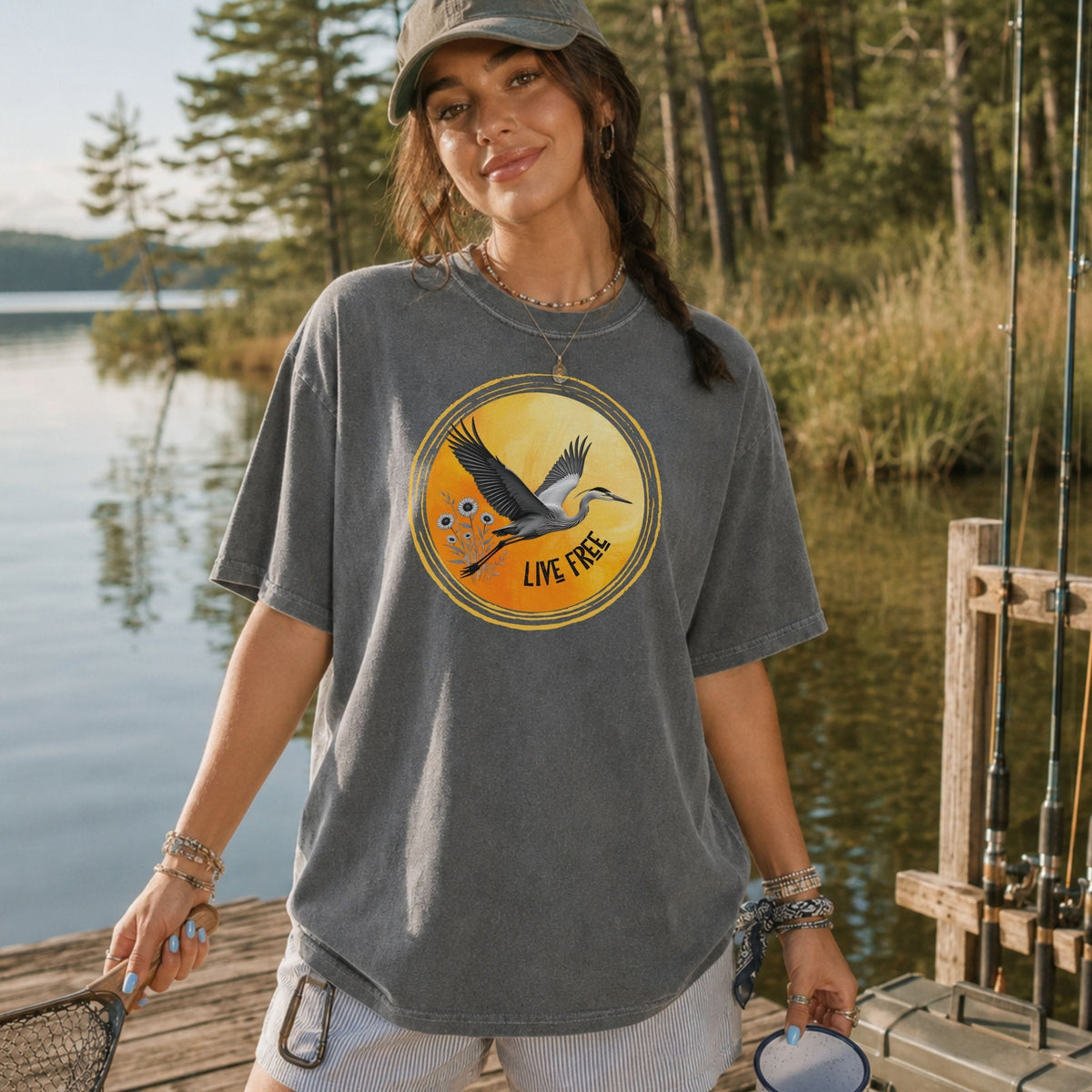 A woman wearing a heather gray graphic t-shirt stands on a wooden dock by a lake. The t-shirt features a circular graphic of a flying bird over orange water with the text 'LIVE FREE'.