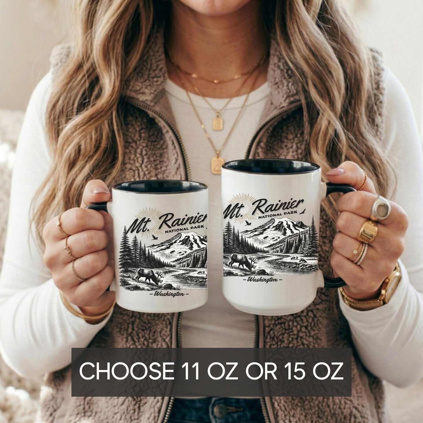 A woman holds two white ceramic mugs with black interiors and handles. The mugs are printed with a black-and-white mountain landscape design and the text 'Mt. Rainier National Park'.