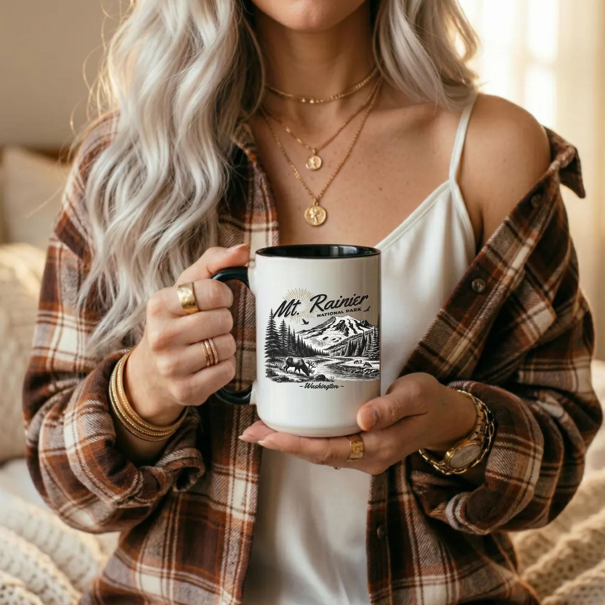 A woman in a brown and beige plaid flannel shirt holds a white ceramic mug with a black handle and rim. The mug features a black line art graphic of Mt. Rainier National Park.