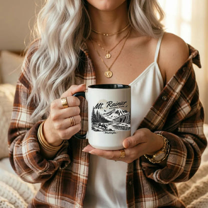 A woman in a brown and beige plaid flannel shirt holds a white ceramic mug with a black handle and rim. The mug features a black line art graphic of Mt. Rainier National Park.