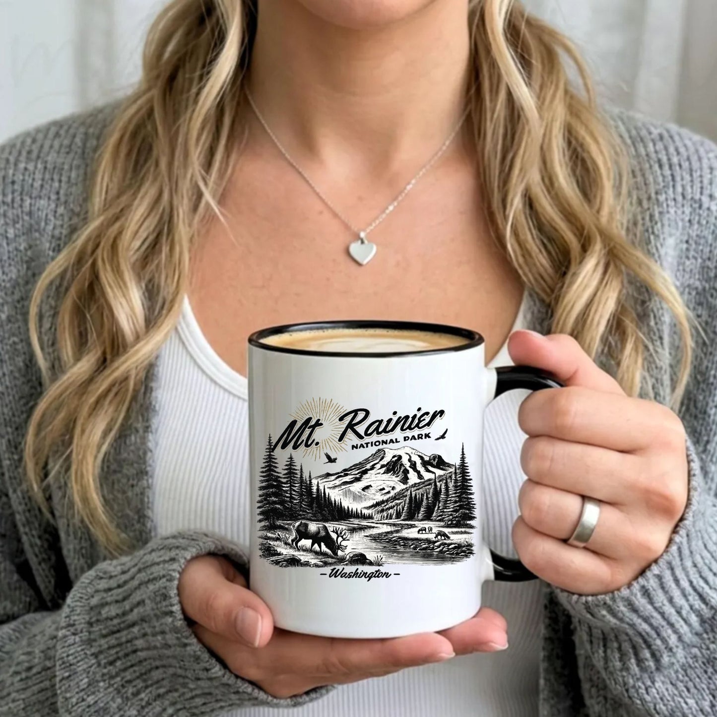 A person holding a white ceramic mug with a black rim, handle, and printed black mountain landscape design labeled 'Mt. Rainier National Park - Washington.