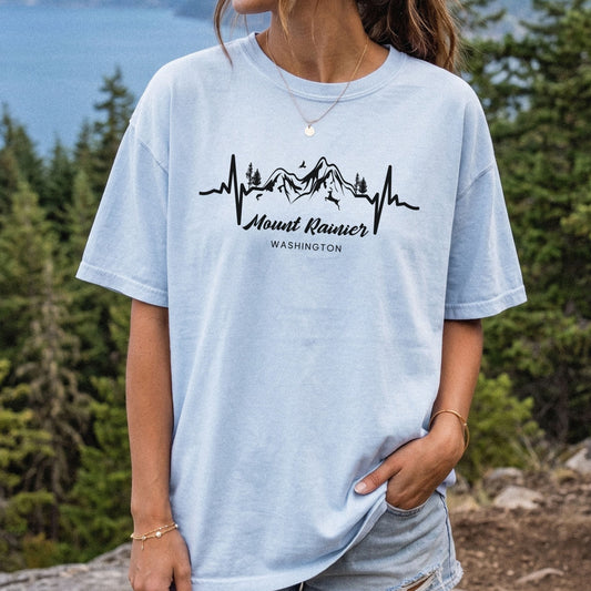 Woman wearing a light blue Mount Rainier Comfort Colors T-shirt with a black mountain heartbeat graphic, standing outdoors on a forested mountain trail in Washington.