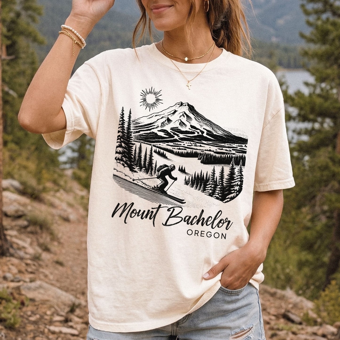 A woman wearing an ivory graphic t-shirt with a black line art print of Mount Bachelor and the text 'Mount Bachelor OREGON'. The background is a vintage style outdoor forest scene.