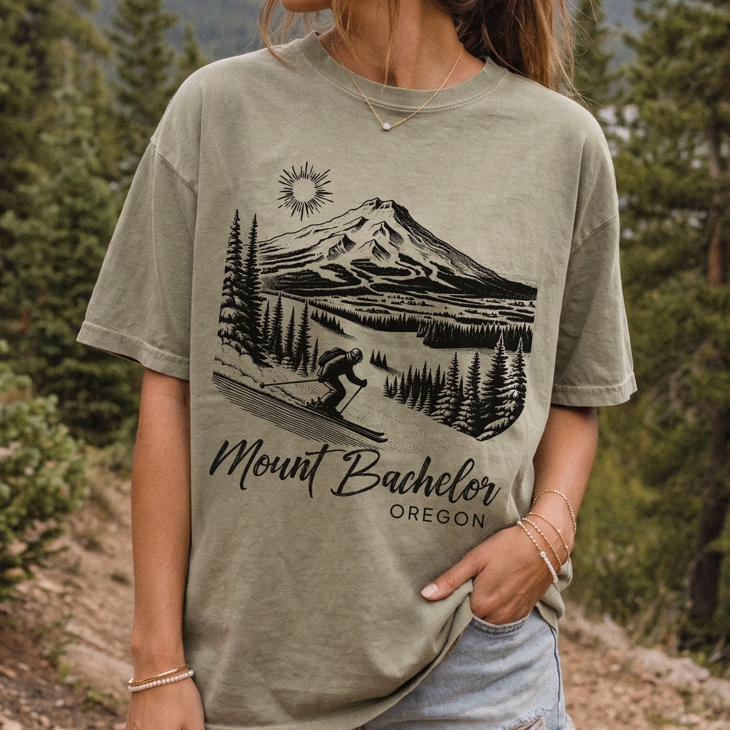 A woman wearing a khaki graphic t-shirt with a black line art print of Mount Bachelor and the text 'Mount Bachelor OREGON'. The background is a vintage style outdoor forest scene.