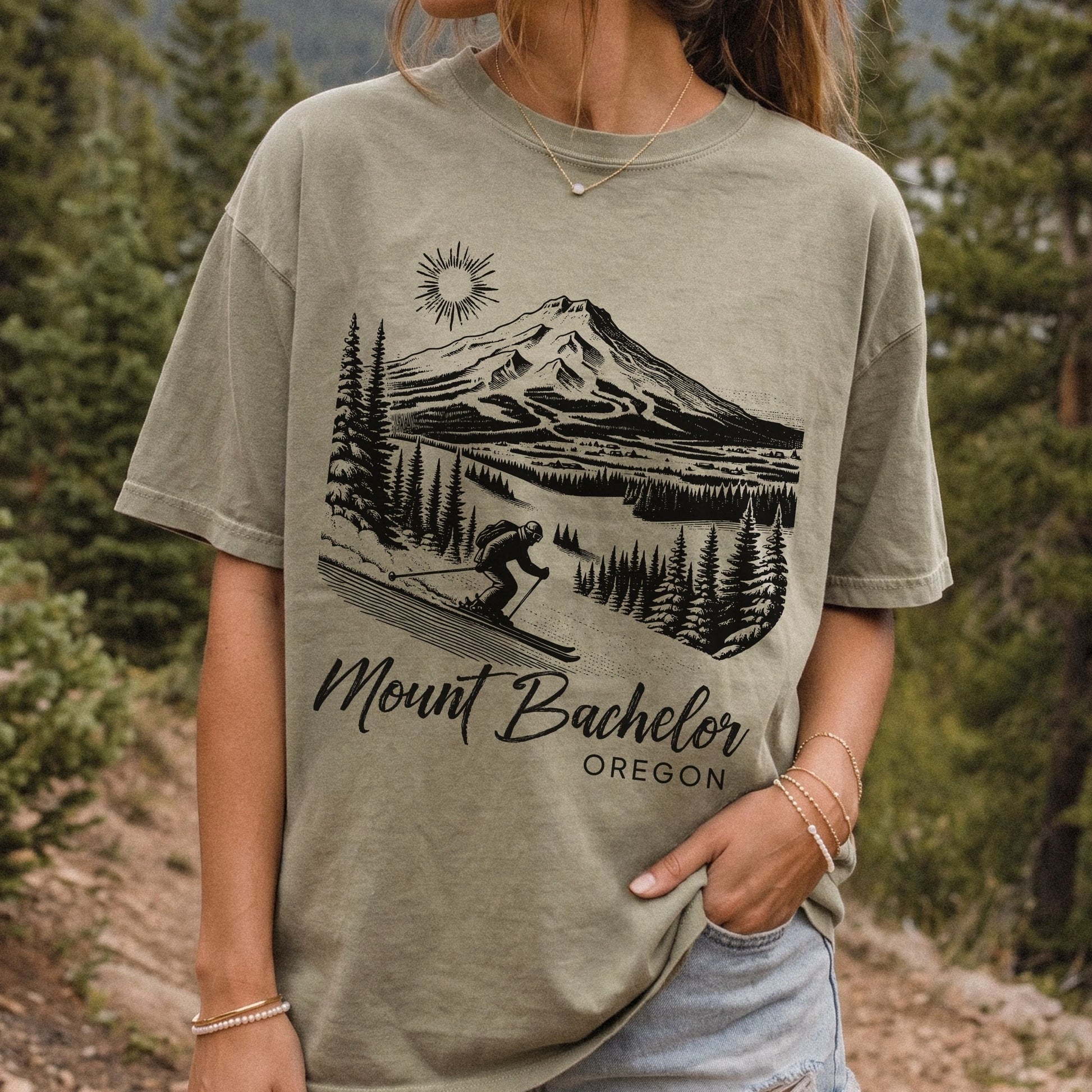 A woman wearing a khaki graphic t-shirt with a black line art print of Mount Bachelor and the text 'Mount Bachelor OREGON'. The background is a vintage style outdoor forest scene.