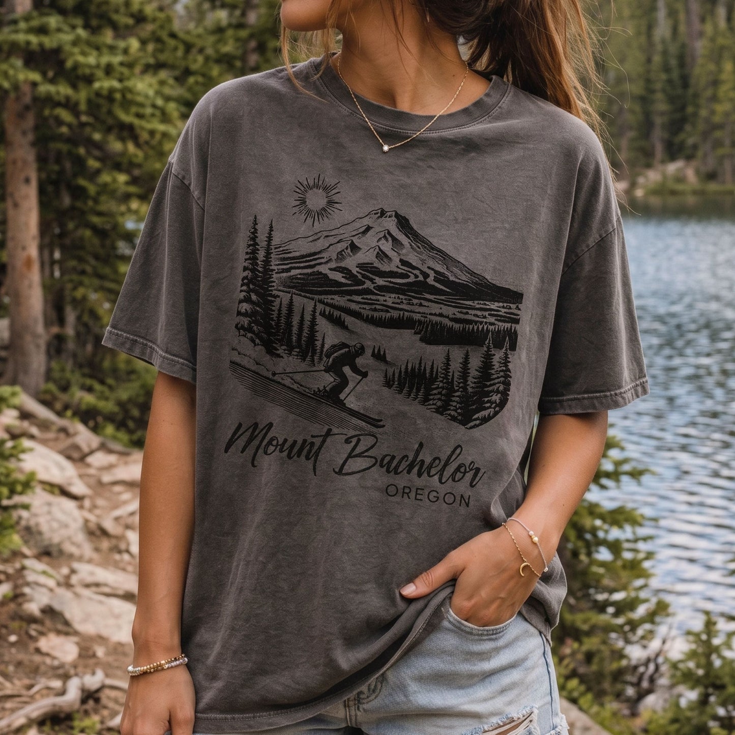 A woman wearing a dark gray mineral-wash graphic t-shirt featuring a black illustration of Mount Bachelor in Oregon with a skier and the text 'Mount Bachelor OREGON'. The background shows a scenic outdoor view of a lake and pine forest.