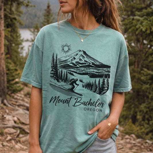 A woman wearing a seafoam green graphic t-shirt with a black line art print of Mount Bachelor and the text 'Mount Bachelor OREGON'. The background is a vintage style outdoor forest scene.