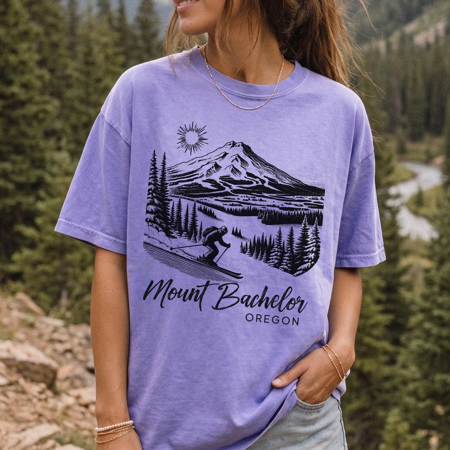 A woman wearing a violet graphic t-shirt with a black line art print of Mount Bachelor and the text 'Mount Bachelor OREGON'. The background is a vintage style outdoor forest scene.