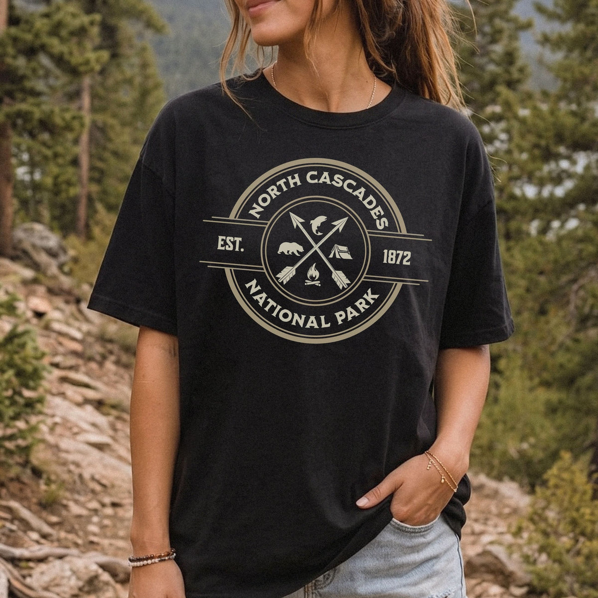 A woman wearing a black graphic t-shirt with a distressed cream-colored circular logo that reads 'North Cascades National Park Est. 1872'.