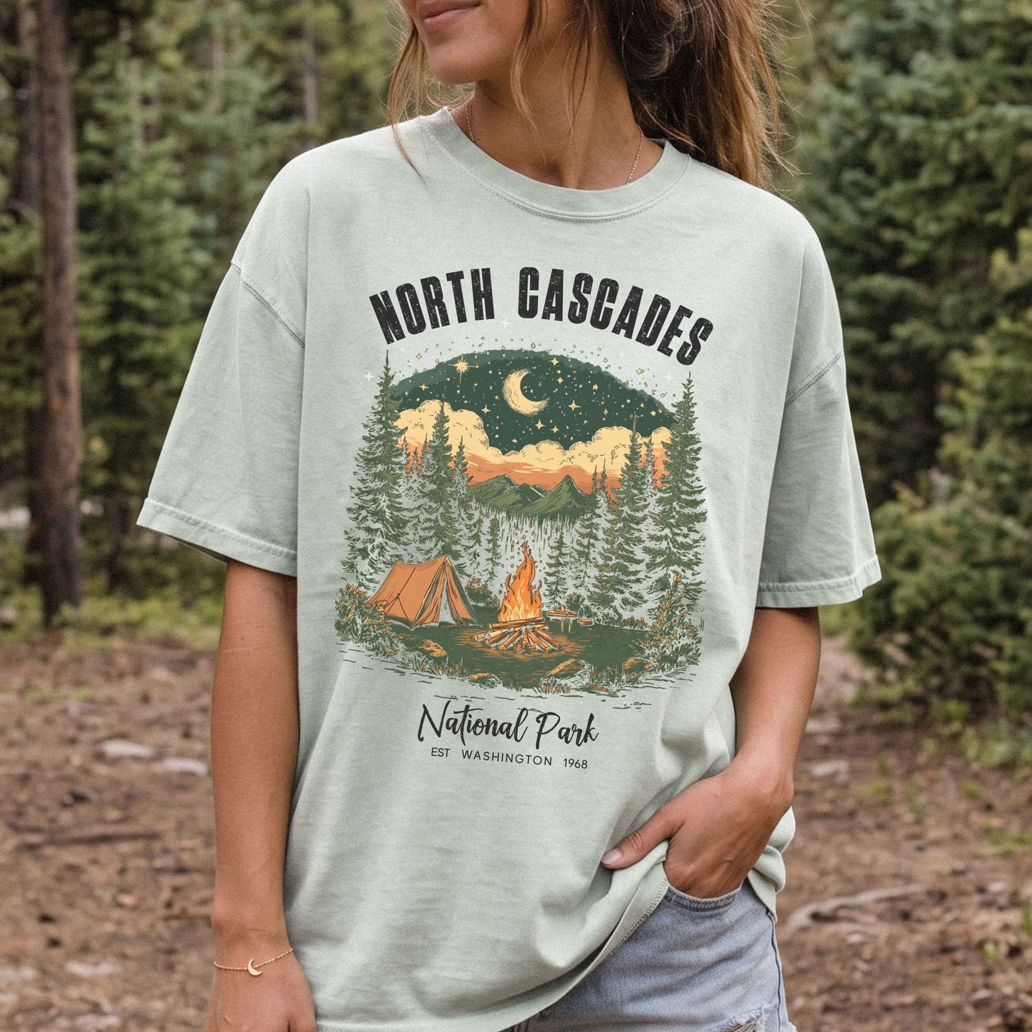 A woman wearing a light sage green graphic t-shirt in an outdoor forest setting. The t-shirt features a printed graphic of a camping scene with a tent, campfire, and mountains under a night sky, with the text 'NORTH CASCADES National Park'.