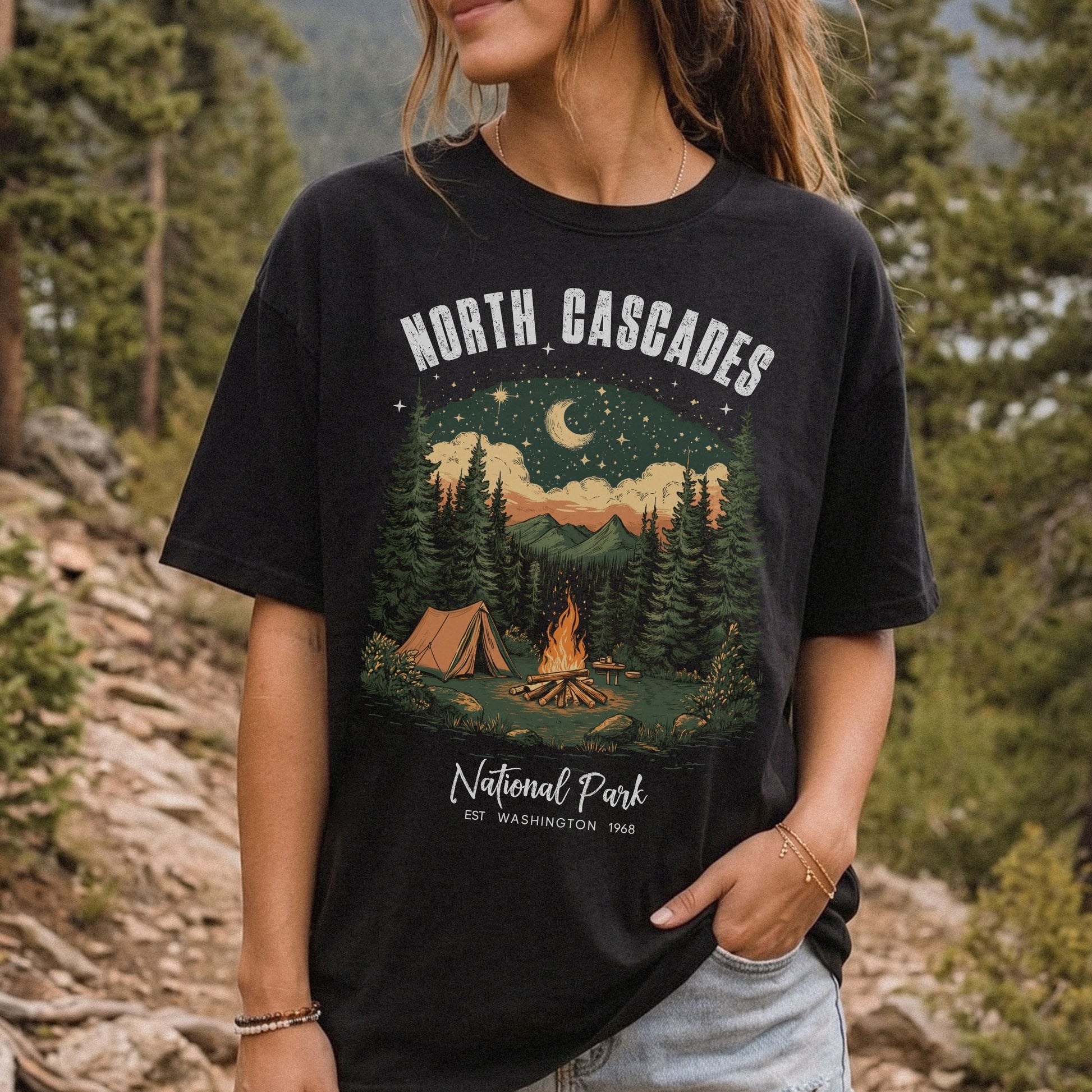 A woman wearing a black graphic t-shirt featuring a 'North Cascades National Park' camping scene, set against a blurred forest background.