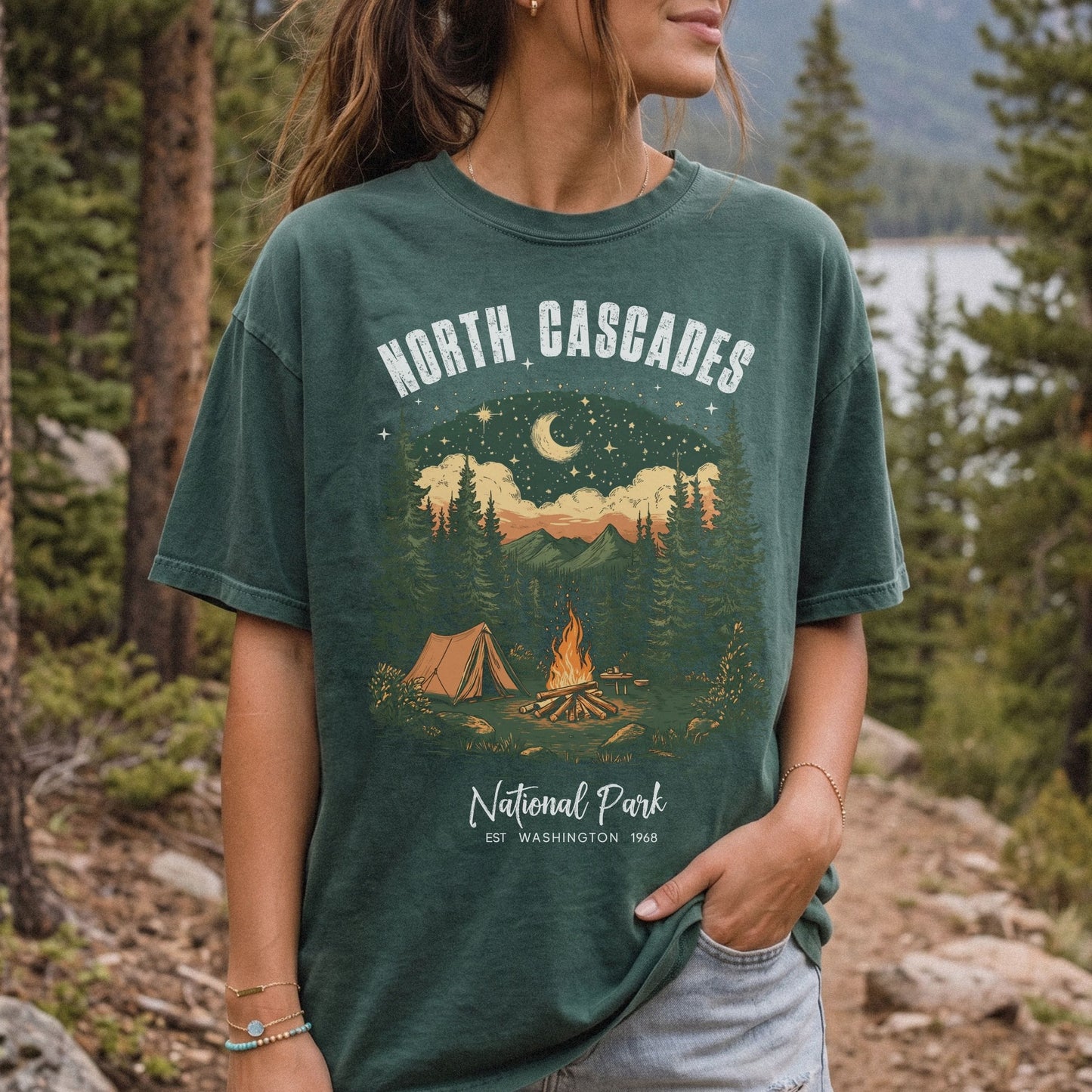 A woman wearing a dark green graphic t-shirt with a 'North Cascades National Park' campsite design, standing outdoors in a forest setting.