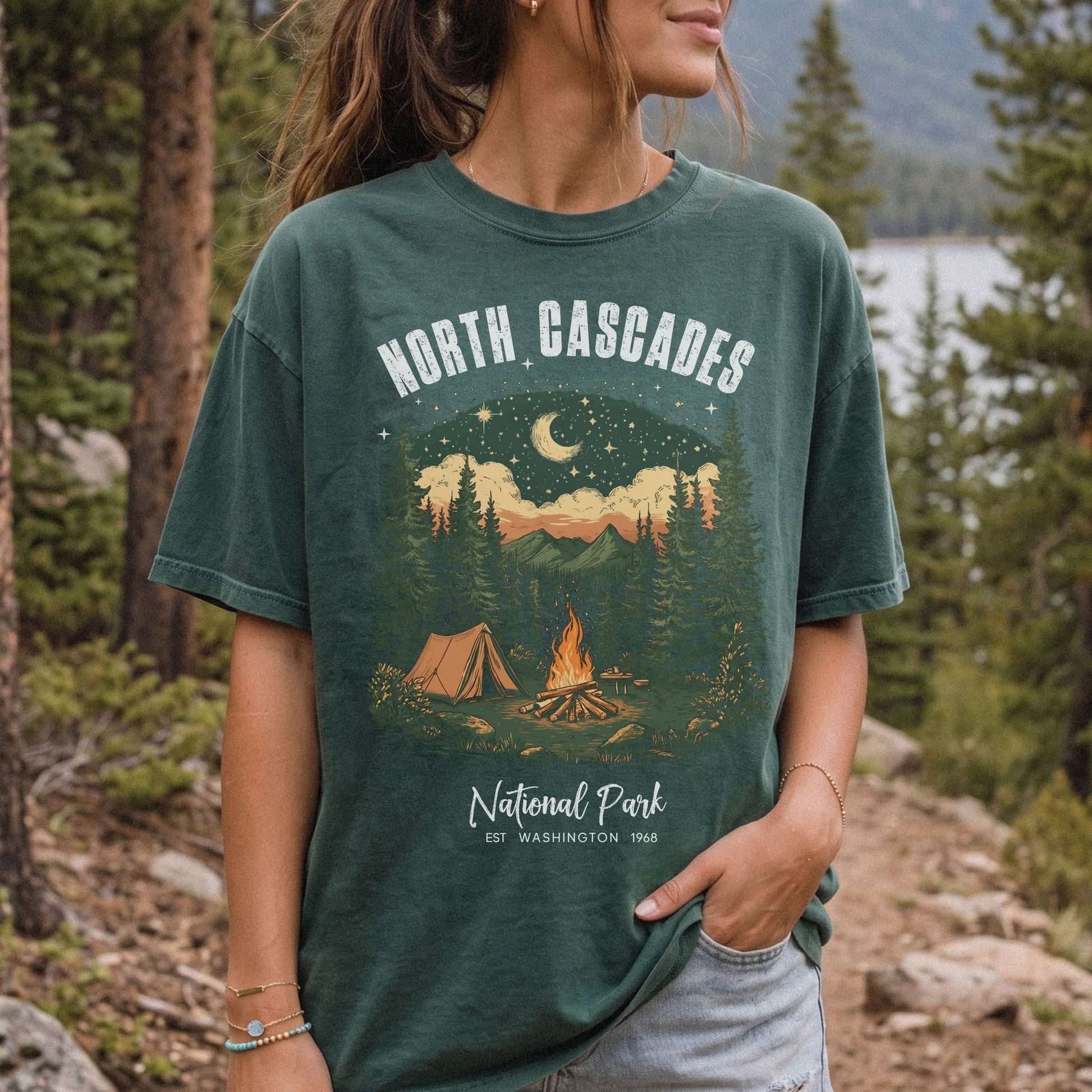 A woman wearing a dark green graphic t-shirt with a 'North Cascades National Park' campsite design, standing outdoors in a forest setting.