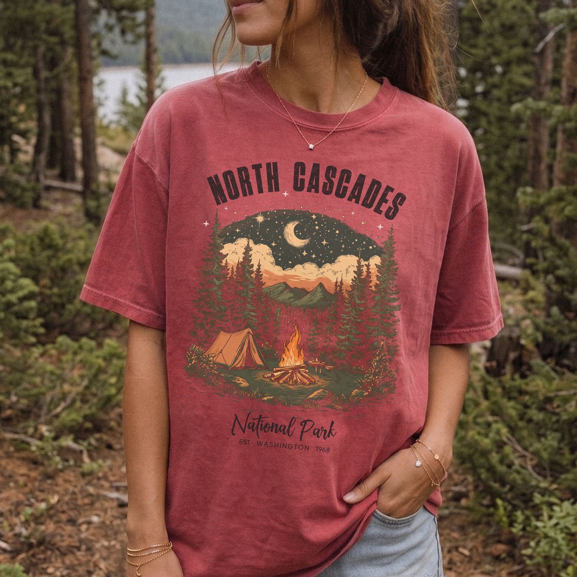 A woman wearing a faded dusty rose t-shirt with a 'North Cascades National Park' camping graphic, standing in a forest setting.