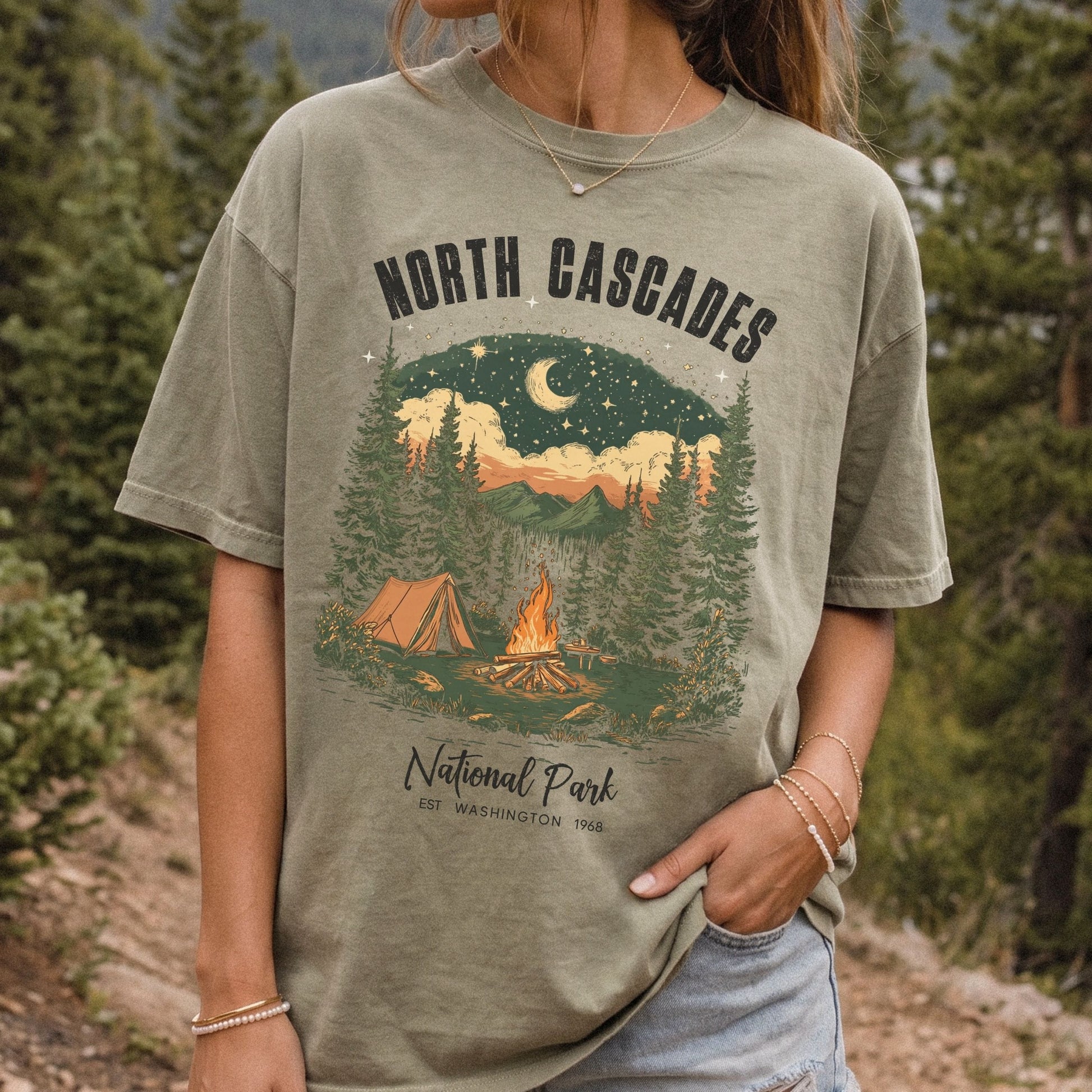 A woman wearing a khaki graphic t-shirt with a 'North Cascades National Park' camping scene printed on the front, set against a blurred forest background.