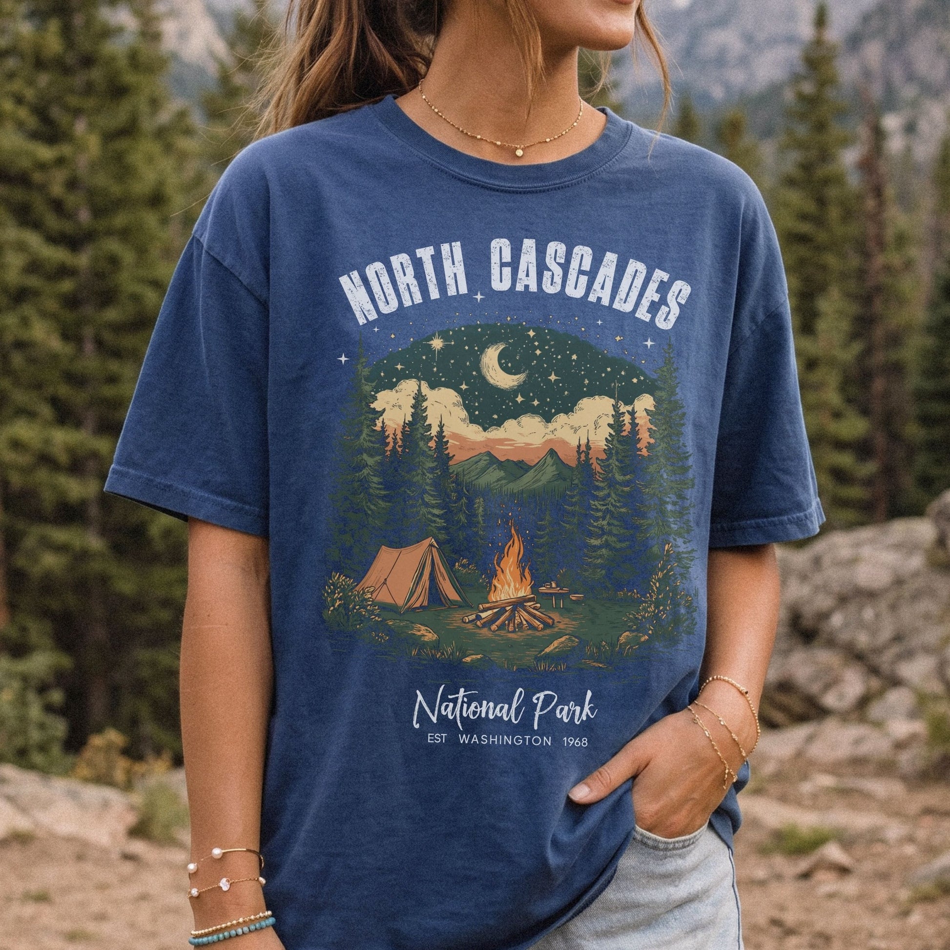 A woman wearing a navy blue graphic t-shirt with a 'North Cascades National Park' camping design, set against a blurred background of pine trees and mountains.