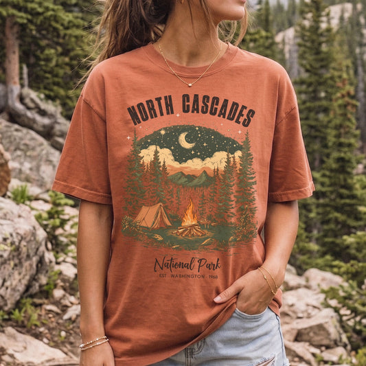 A woman wearing a rust-orange graphic t-shirt with a 'North Cascades National Park' camping scene print, set against a blurred forest and mountain background.