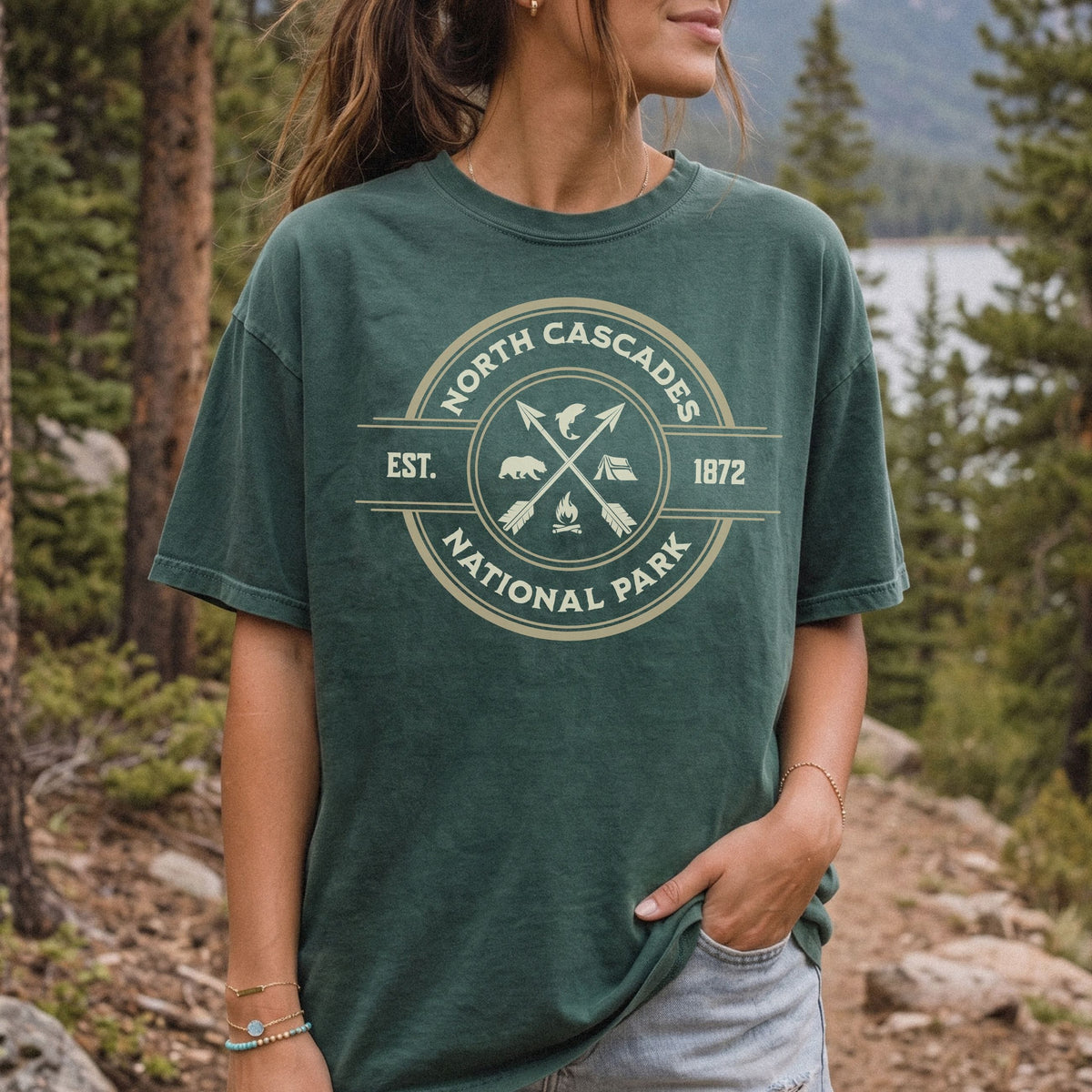 A woman wearing a dark green graphic t-shirt with a vintage-style circular logo for North Cascade National Park, set against a natural forest background.