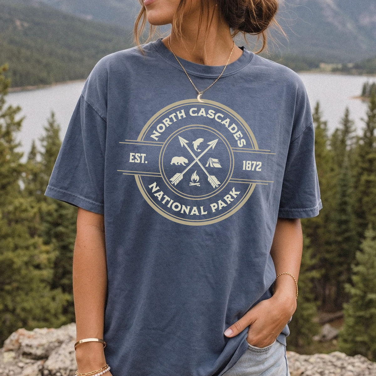 A woman wearing a faded slate blue graphic t-shirt with a cream-colored circular 'North Cascades National Park' logo, set against a scenic outdoor background of a lake and pine trees.