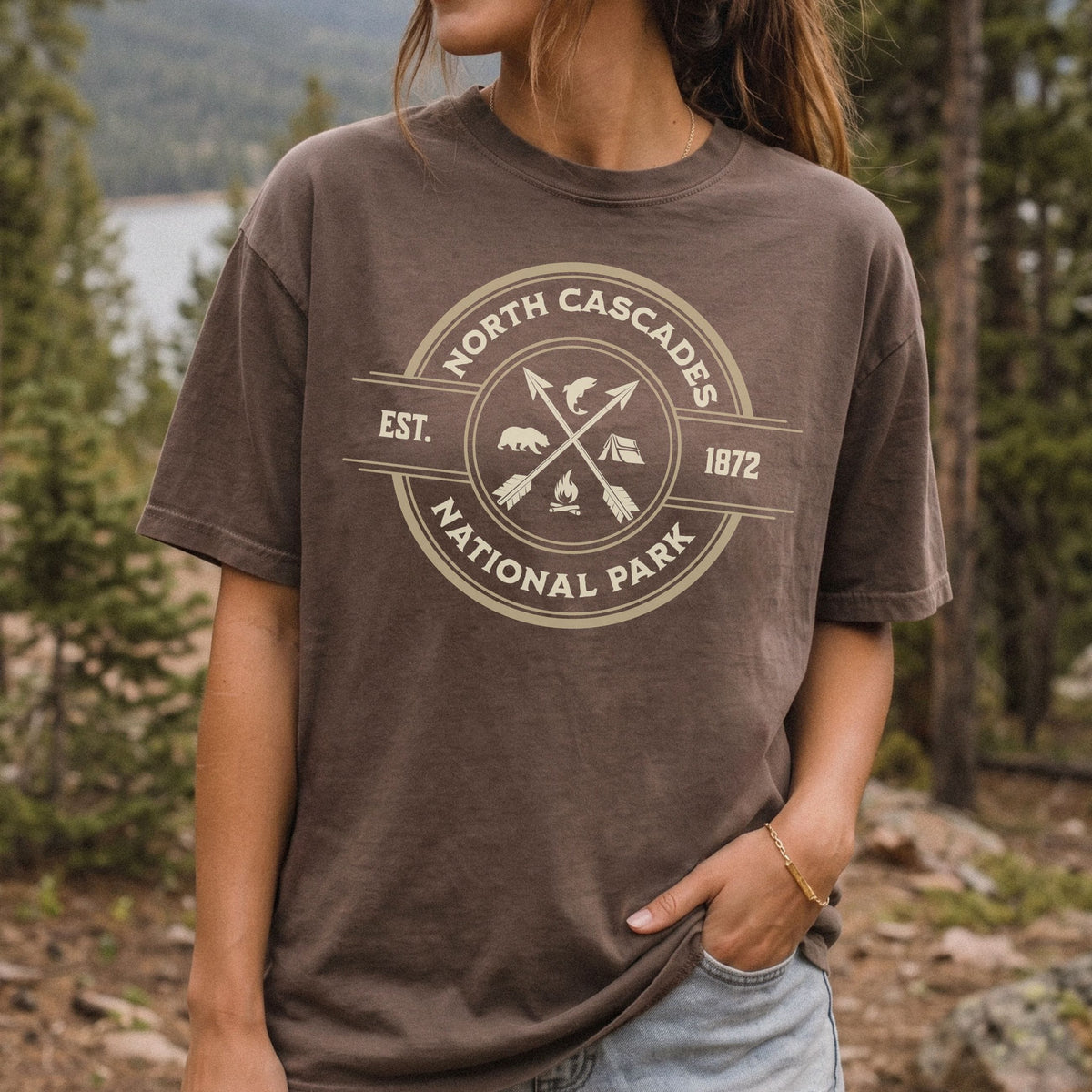 A woman wearing a brown t-shirt with a beige circular graphic that reads 'NORTH CASCADES NATIONAL PARK EST. 1872' with crossed arrows and campfire icons, set against a blurred forest background.