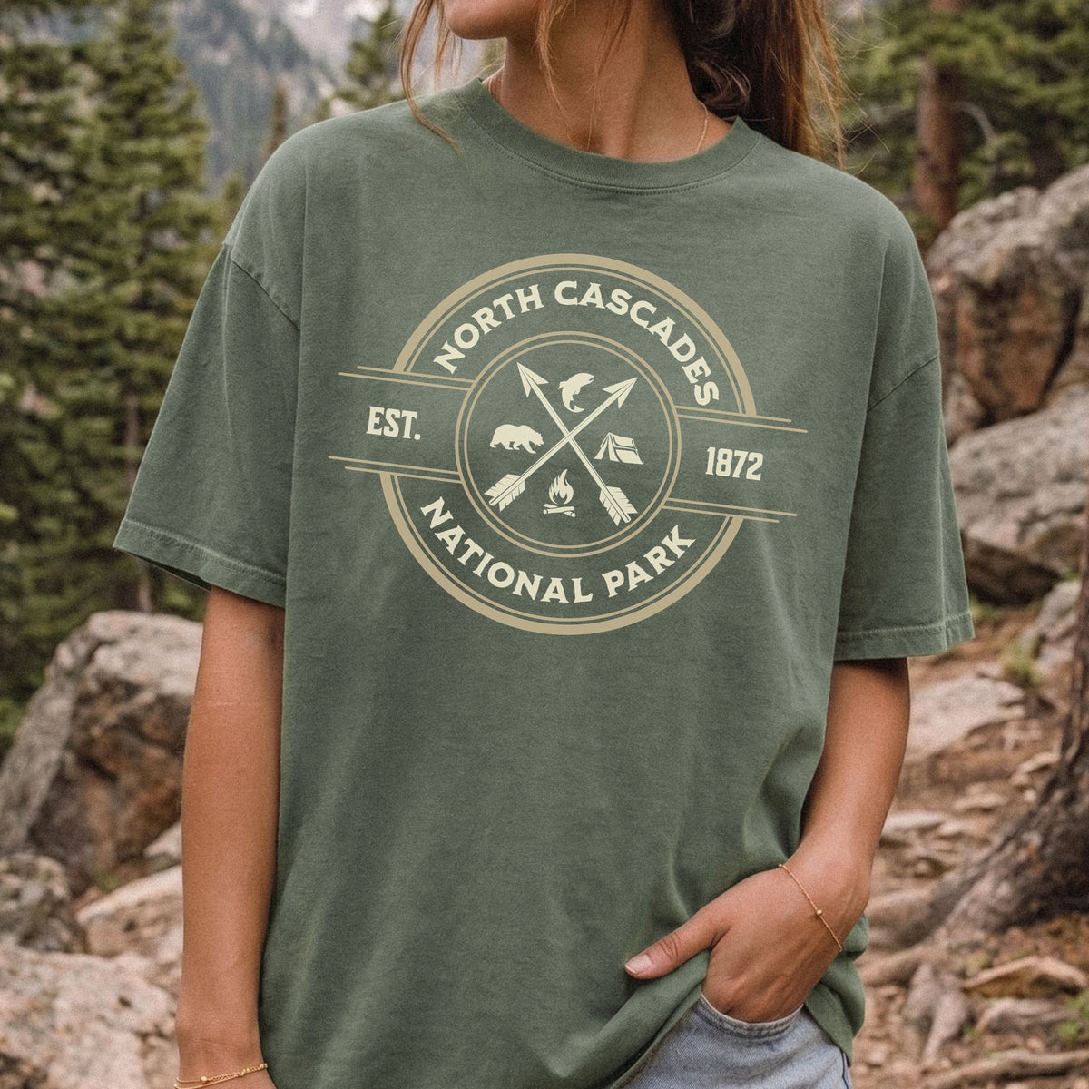 A woman wearing an oversized olive green t-shirt with a cream-colored circular graphic that reads 'NORTH CASCADES NATIONAL PARK EST. 1872' with crossed arrows, campfire, and bears.