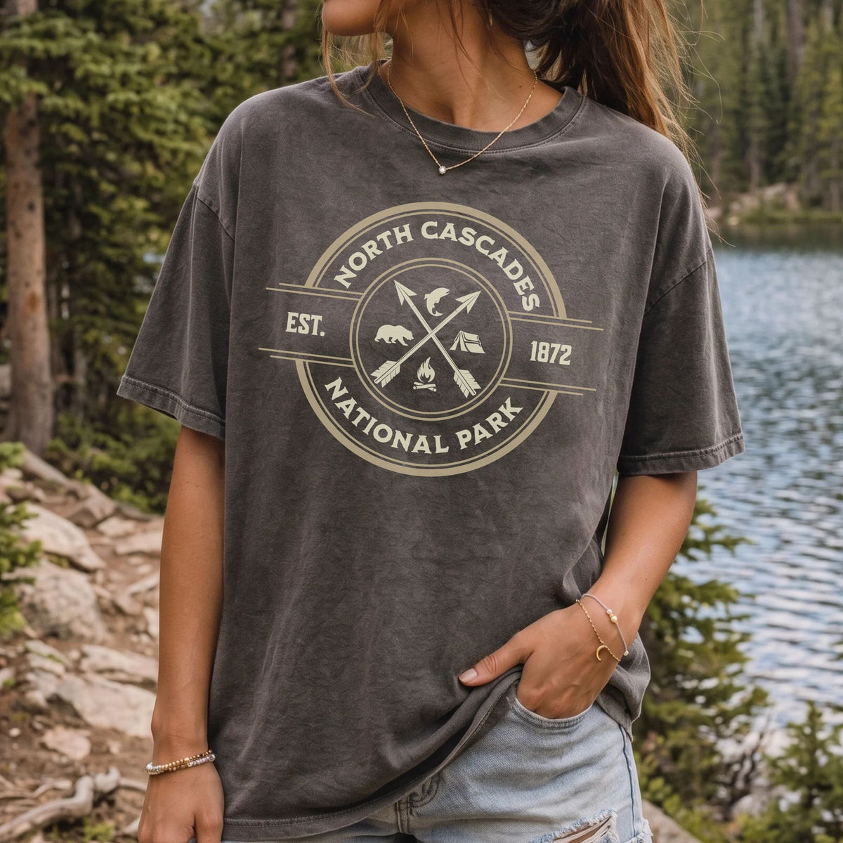 A woman wearing a dark gray acid-wash graphic t-shirt with a cream-colored circular 'North Cascades National Park' print, standing outdoors near a lake and forest.