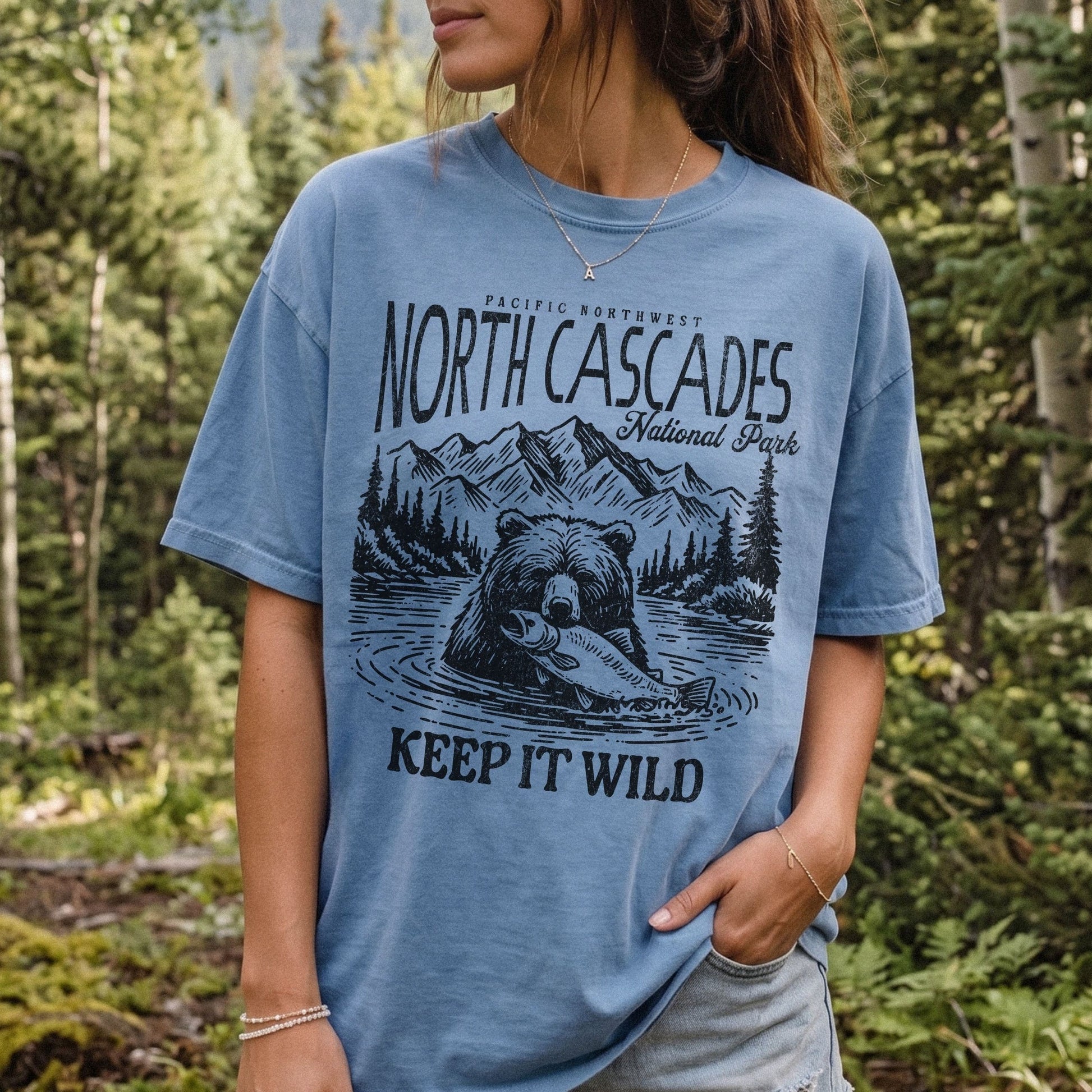 A woman wearing a light blue graphic t-shirt with a black print of a bear, mountains, and the text 'NORTH CASCADES National Park' and 'KEEP IT WILD', photographed outdoors in a forest setting.