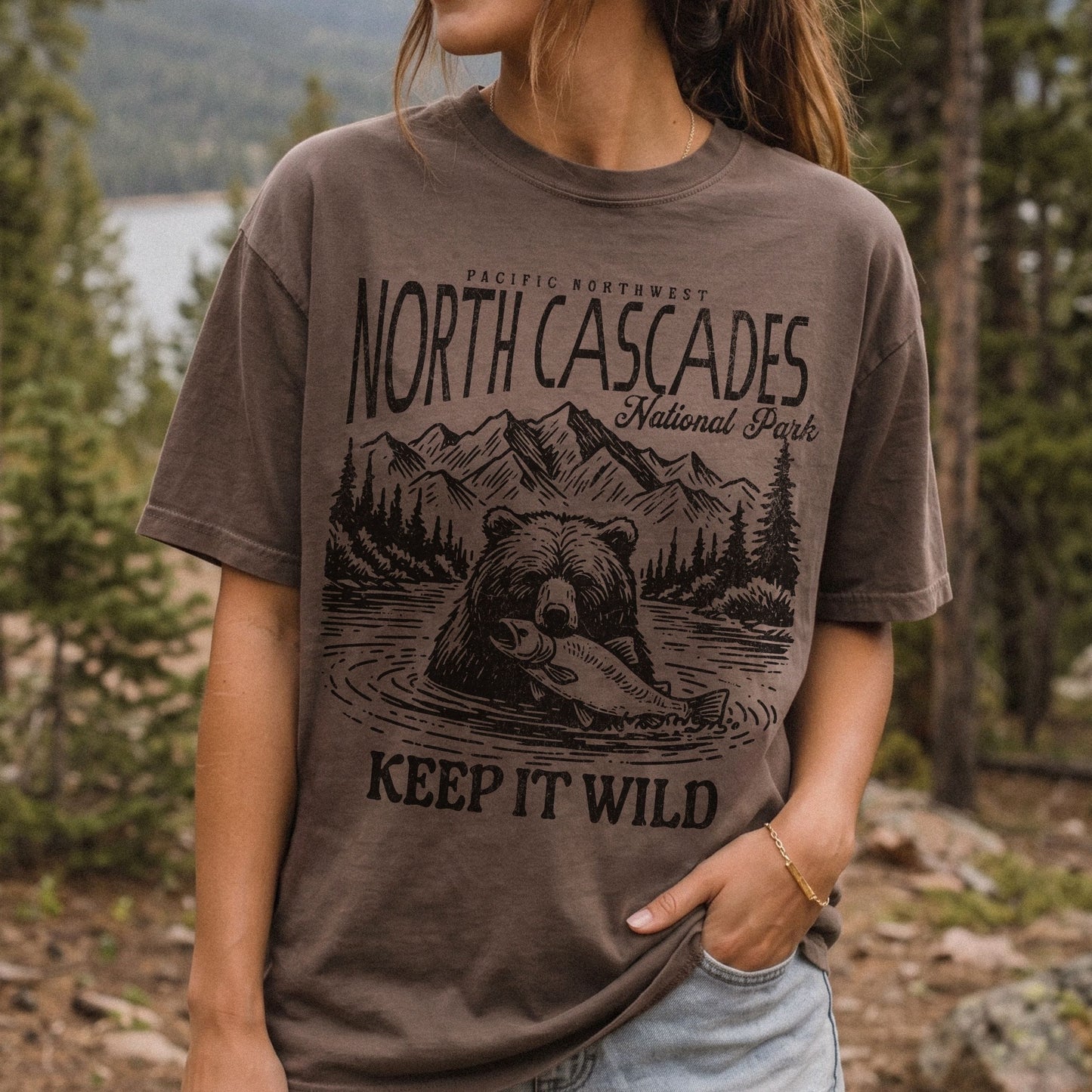 A woman wearing a brown graphic t-shirt with a black print of a bear, mountains, and the text 'NORTH CASCADES National Park' and 'KEEP IT WILD', set against a blurred forest background.