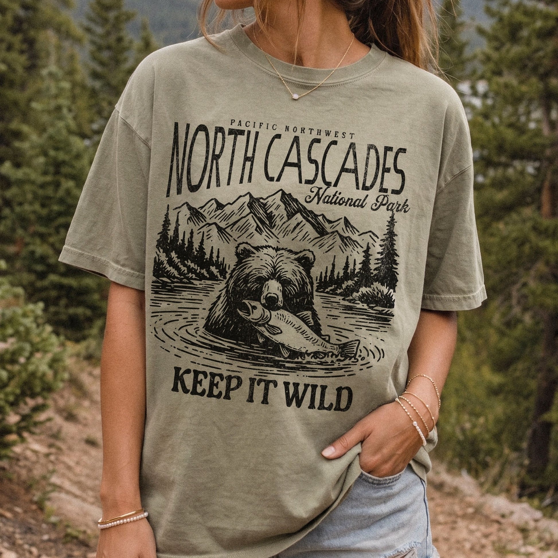 A woman wearing an oversizedkhaki graphic t-shirt featuring a black print of a bear fishing in a river with mountains and the text 'NORTH CASCADES National Park' and 'KEEP IT WILD', set against a blurred forest background.