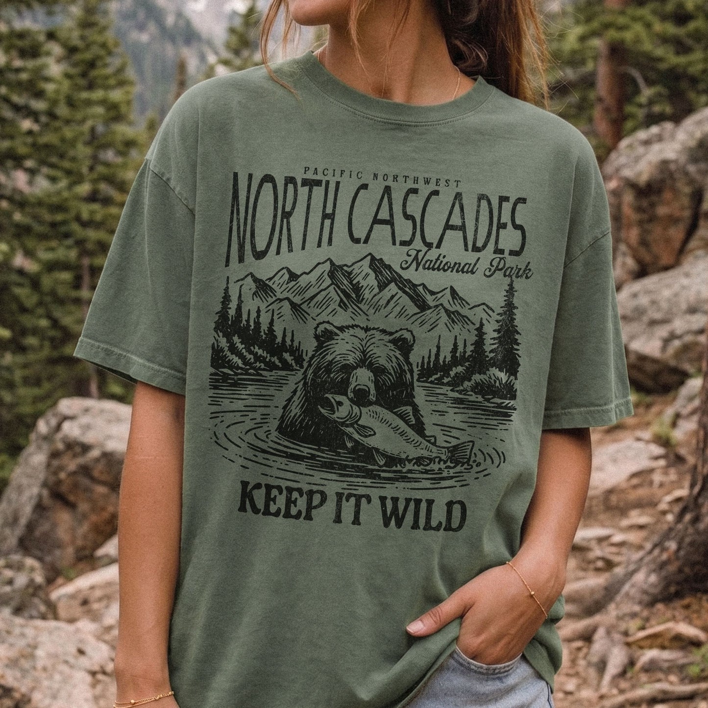A woman wearing an oversized moss green graphic t-shirt with a black print of a grizzly bear holding a salmon, mountains, and the text 'NORTH CASCADES National Park' and 'KEEP IT WILD', set against a blurred outdoor forest and rock background.