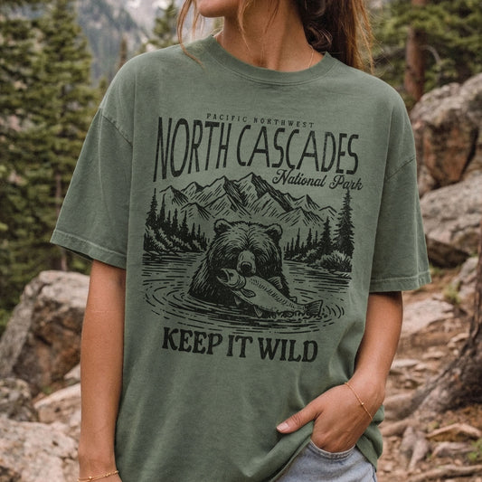 A woman wearing an oversized moss green graphic t-shirt with a black print of a grizzly bear holding a salmon, mountains, and the text 'NORTH CASCADES National Park' and 'KEEP IT WILD', set against a blurred outdoor forest and rock background.