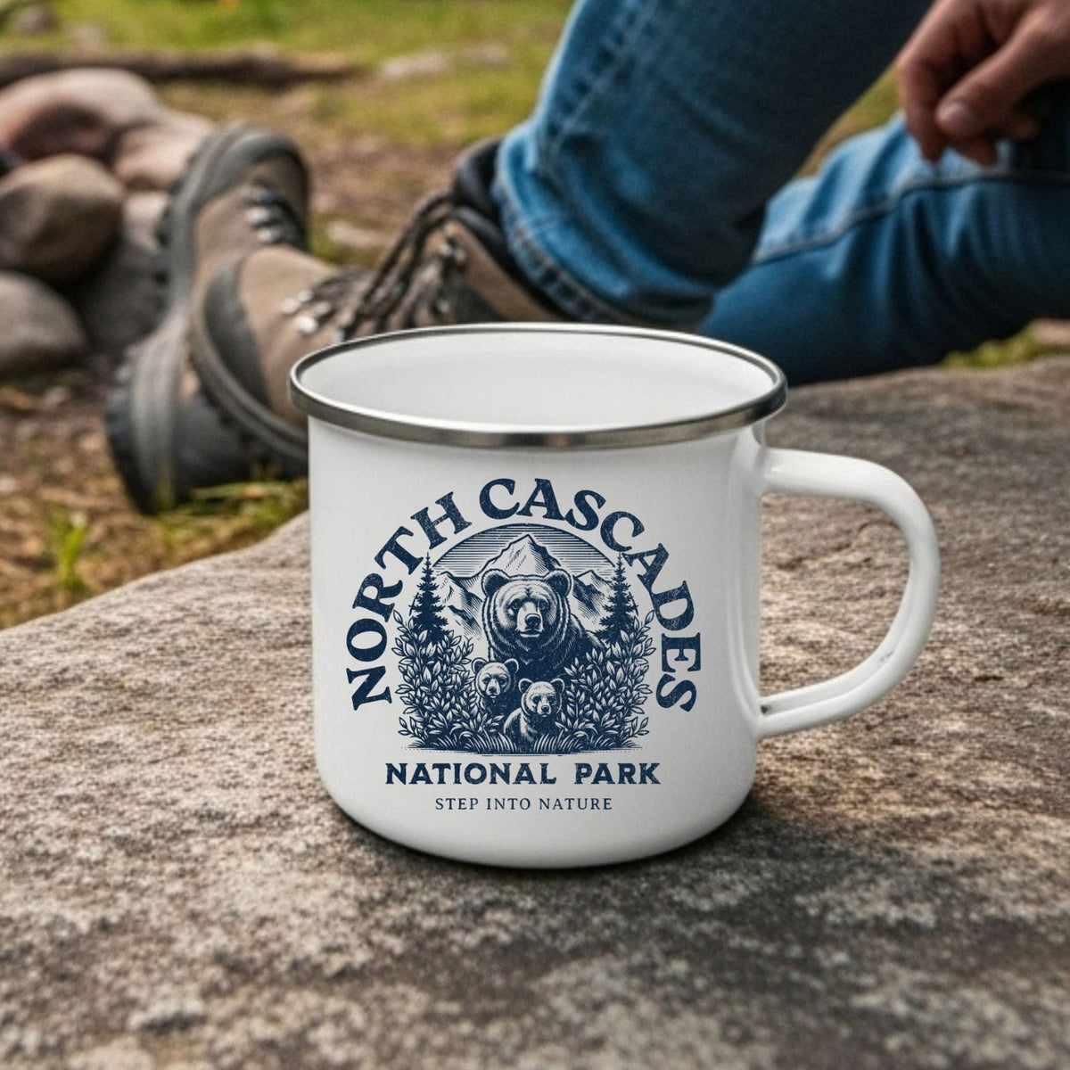 A white enamel camping mug with a blue 'North Cascade National Park' graphic sits on a large stone in the foreground. In the background, a person wearing blue jeans and hiking boots sits near a campfire with an orange tent visible in the distance, creating a natural outdoor setting.