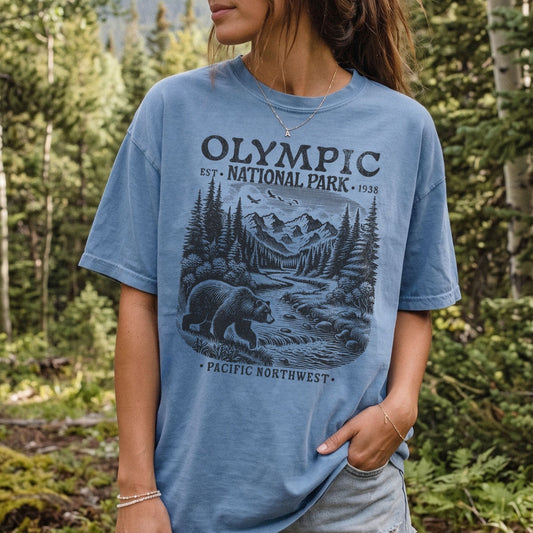 A woman wearing a light blue graphic t-shirt with a black and gray 'Olympic National Park' bear and mountain design, set against a blurred forest background.