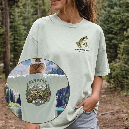 A woman wearing a light sage green graphic t-shirt in an outdoor forest setting. The t-shirt features a green tree frog and 'Olympic National Park' text on the left chest, and a large matching graphic on the back.