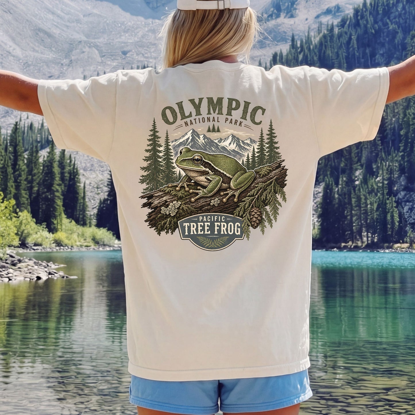 A person standing outdoors in front of a lake and mountains, wearing a cream T-shirt with a large back graphic that reads 'Olympic National Park' and 'Pacific Tree Frog' with an illustration of a frog, trees, and mountains. The person has their arms outstretched and is wearing blue shorts.