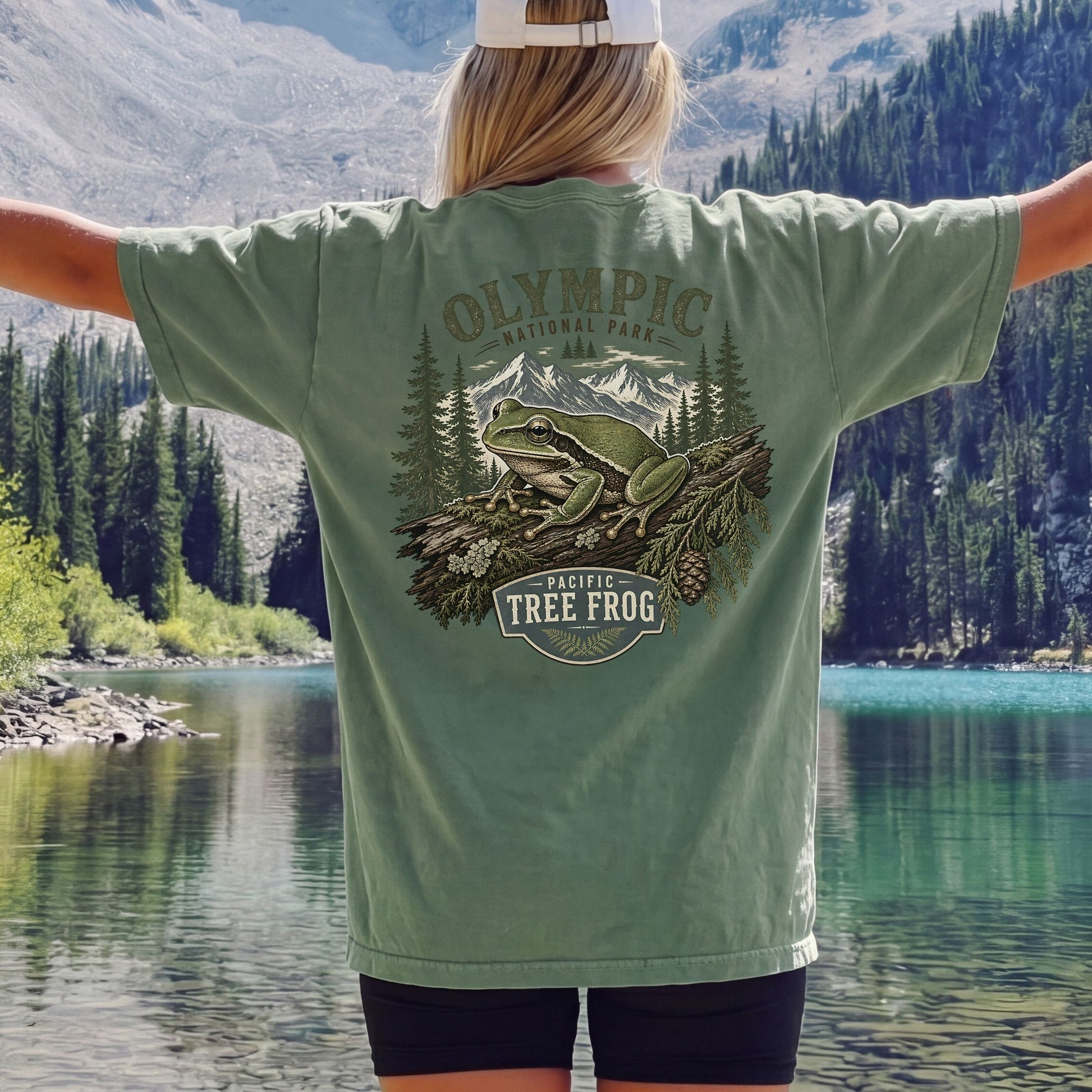 A person standing outdoors by a lake with mountains and pine trees in the background, wearing an oversized sage green graphic T-shirt. The back of the shirt features a large printed design with a frog, mountains, trees, and the text 'OLYMPIC National Park' and 'PACIFIC TREE FROG'.