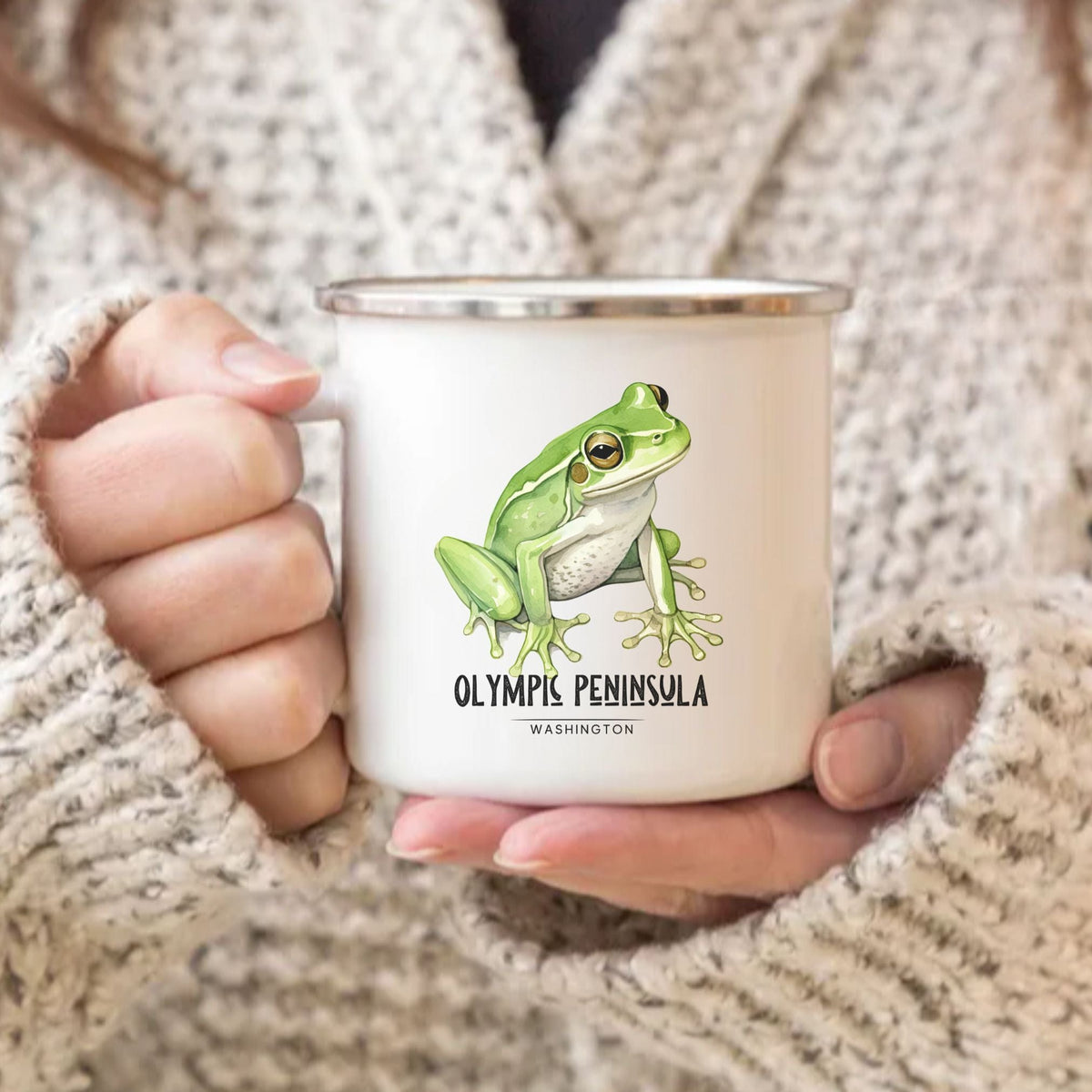 Person holding a white enamel camp mug featuring a green frog illustration and the words “Olympic Peninsula Washington.”