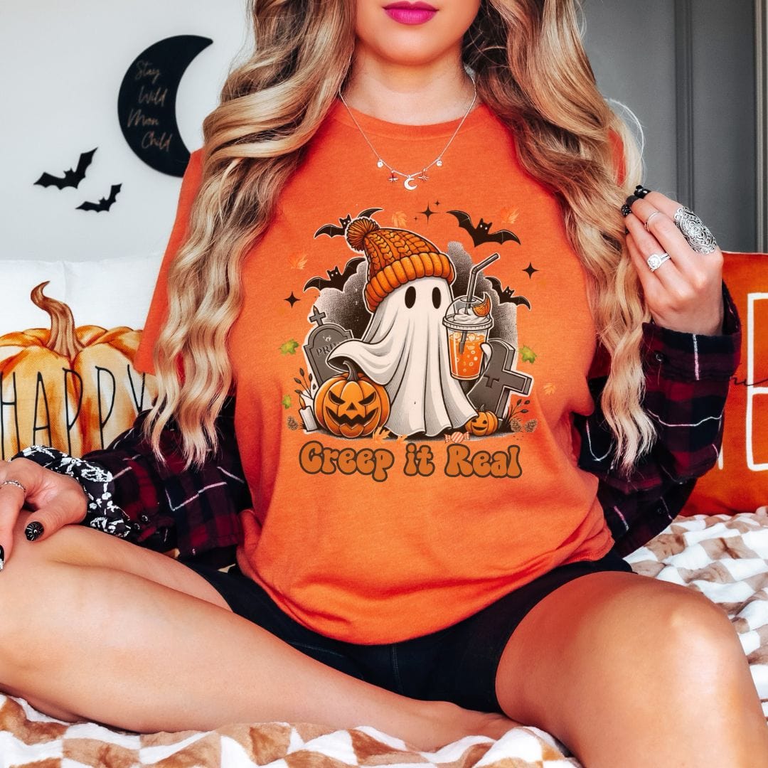 A person with long blonde hair and a black and red plaid shirt sits with their legs crossed on a checkered bed, wearing an orange graphic tee. The shirt features a ghost wearing an orange knit beanie, holding an iced drink, and surrounded by bats, jack-o-lanterns, and text that says, 'Creep it Real.'